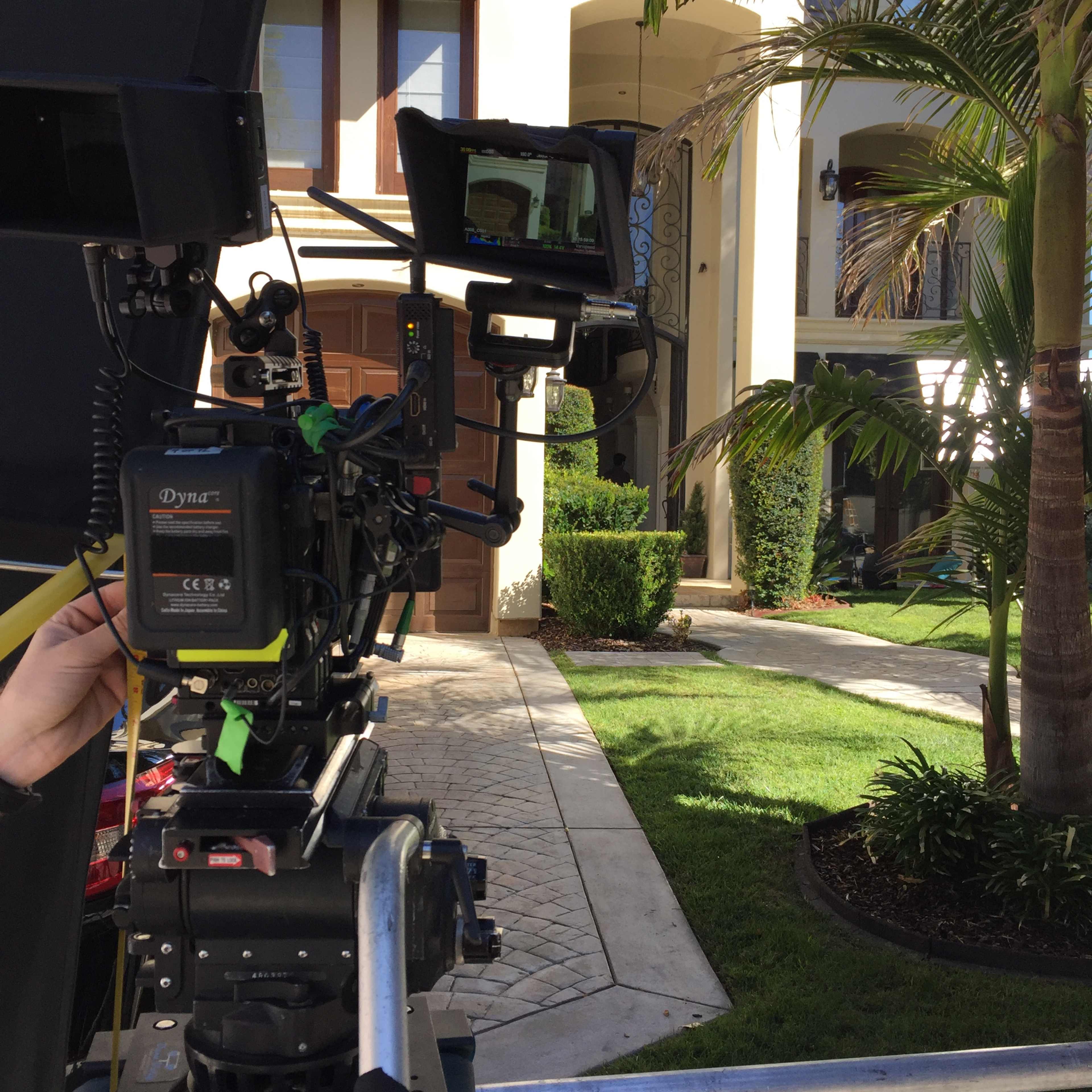 A camera setup is positioned in front of a large house, with palm trees and manicured landscaping visible in the background.
