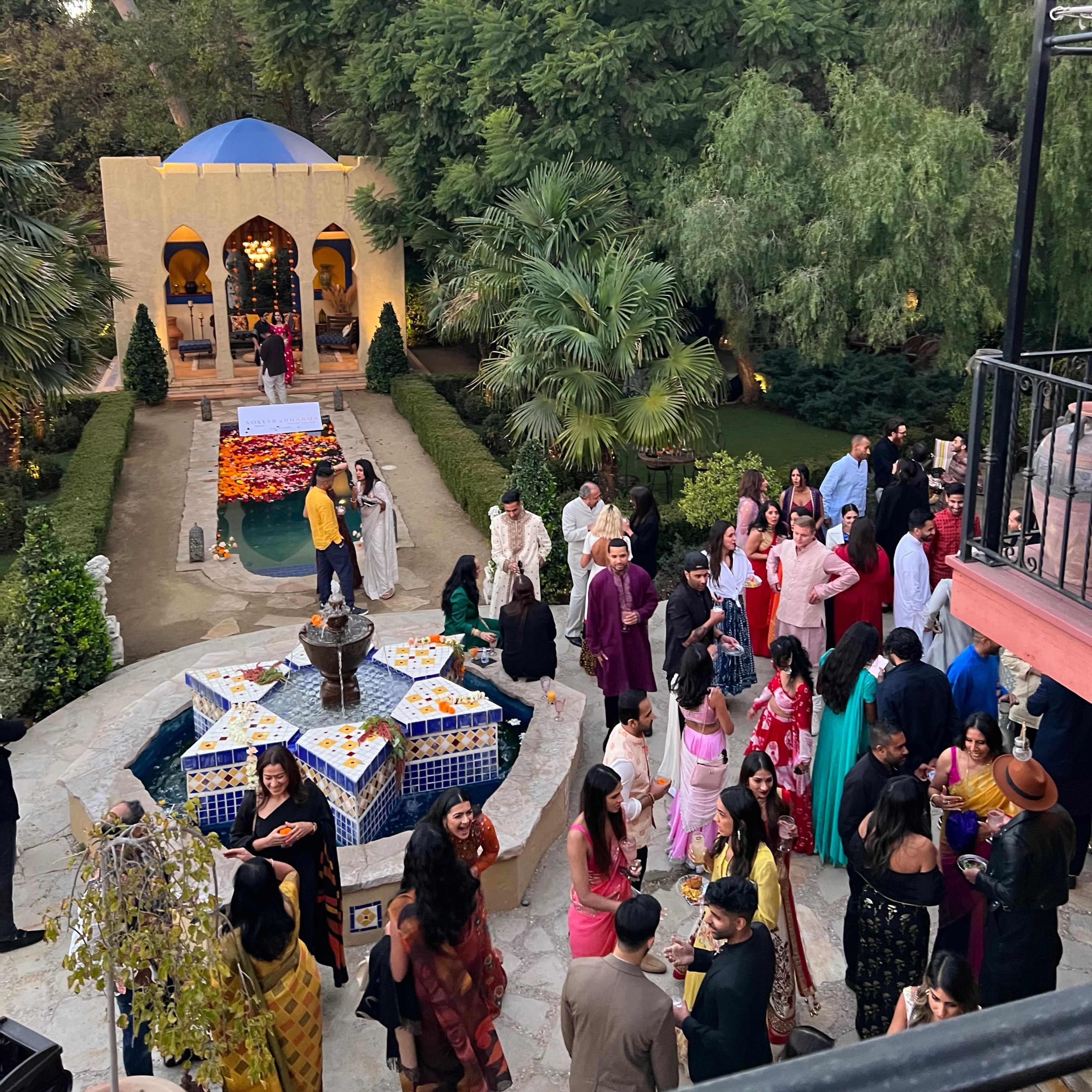 A vibrant outdoor gathering takes place in a lush garden, featuring guests in traditional attire around a decorated fountain and a table of colorful decorations.
