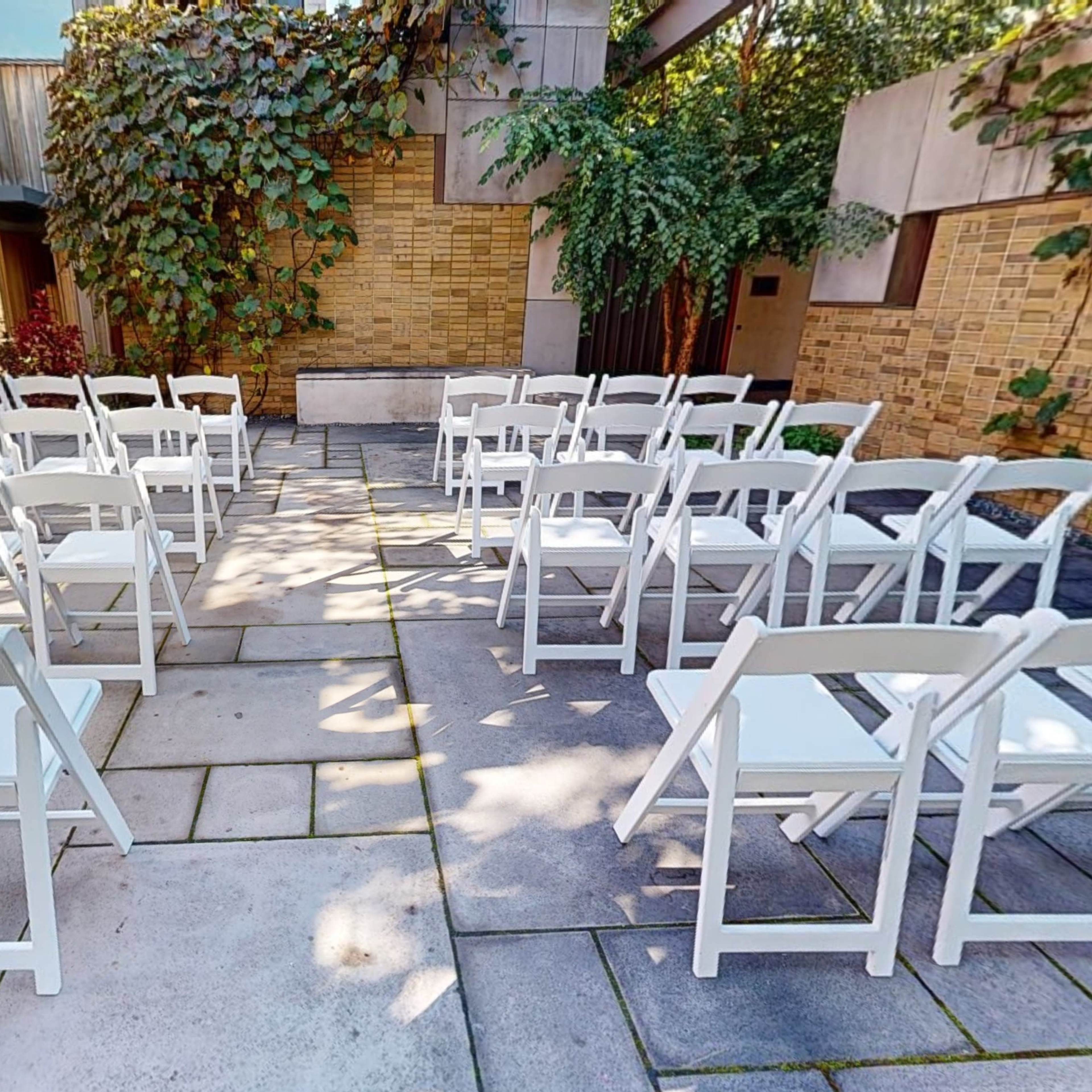 A courtyard arranged with white folding chairs facing a central area, surrounded by brick walls and greenery.