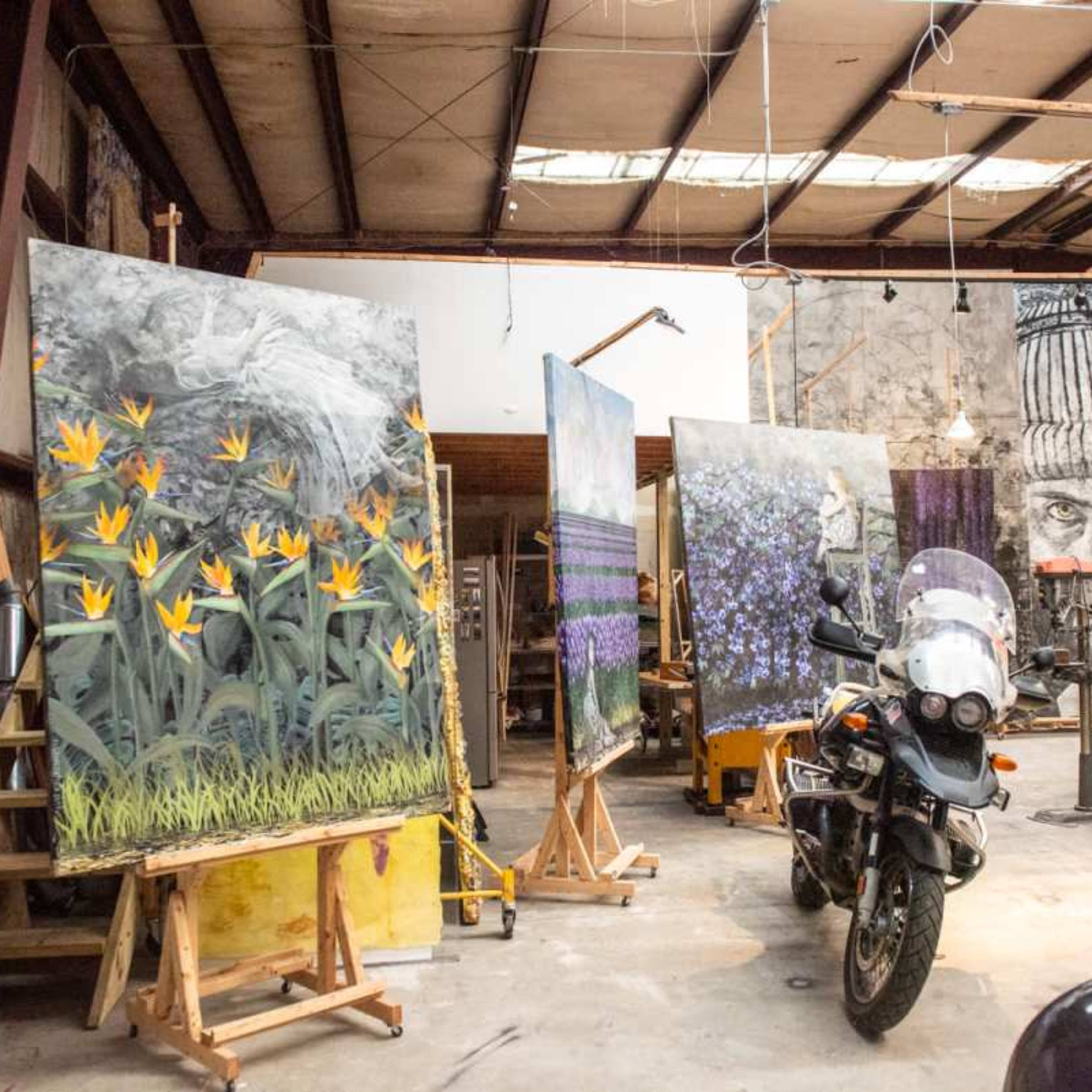 An art studio with several large paintings on easels, a motorcycle parked in the foreground, and various artworks displayed around the room.