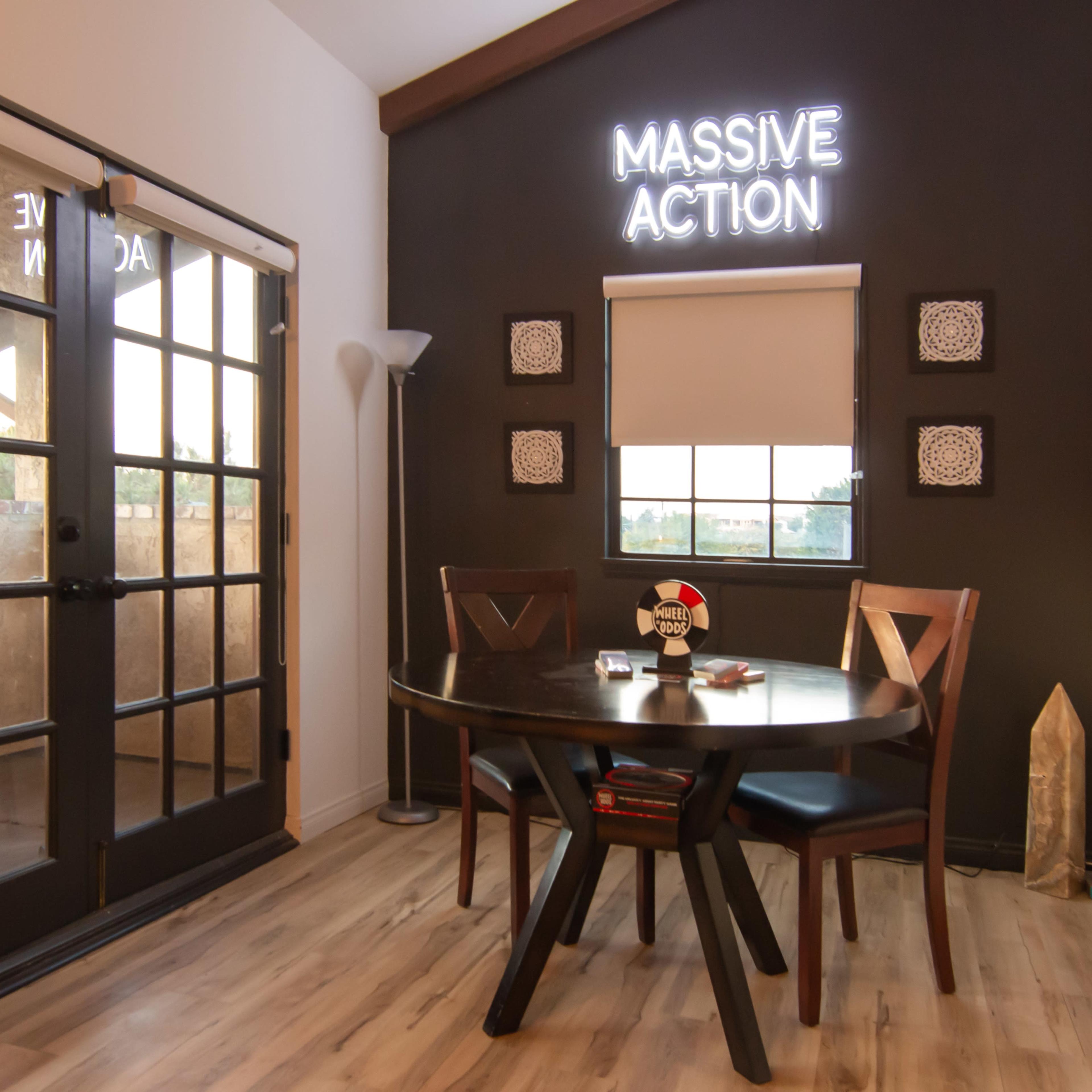 A small meeting room features a round table with two chairs, a large glass door, and a wall displaying the phrase "MASSIVE ACTION" in illuminated letters.