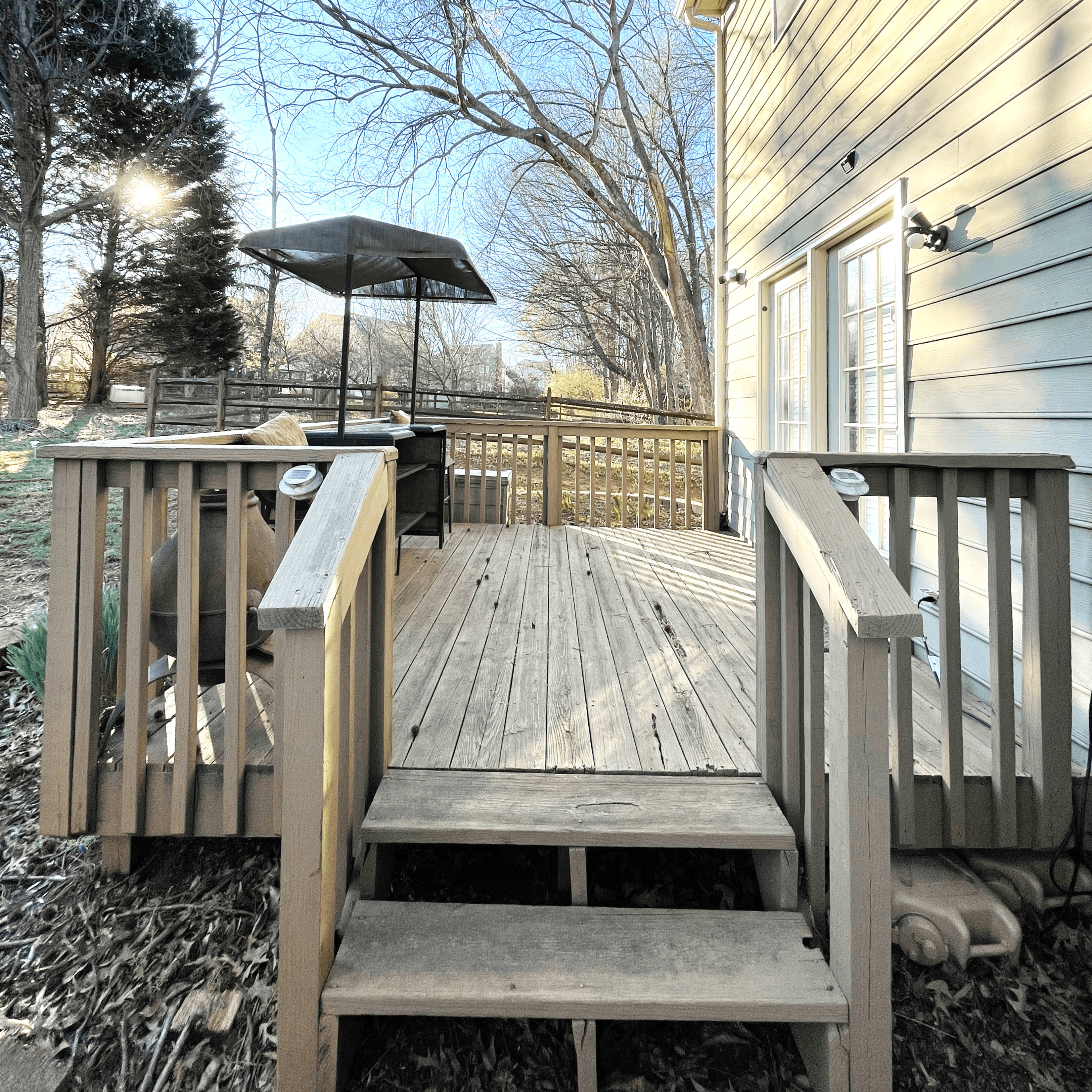 A wooden deck with steps leads to a house, surrounded by bare trees and a patio with an umbrella in the background.