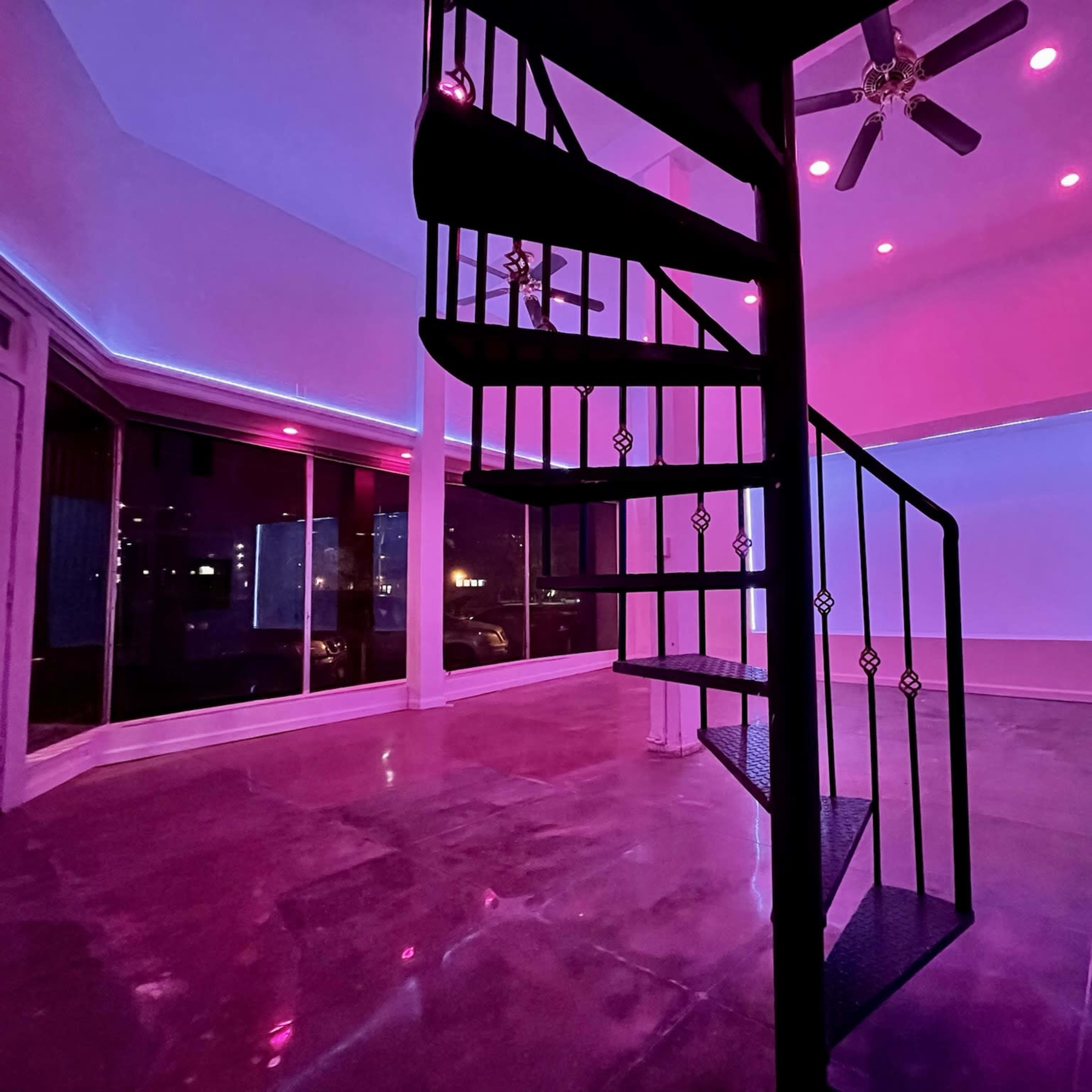 A spiral staircase is centered in a room with large windows that emit pink and purple lighting.