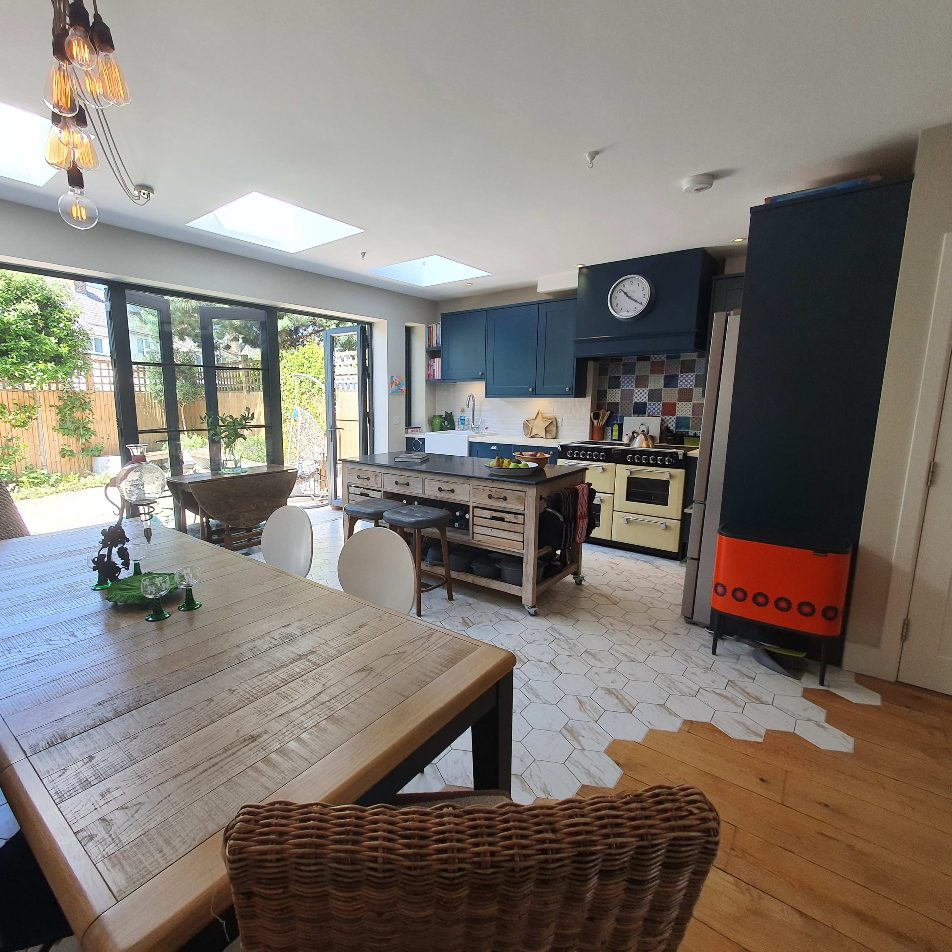 A modern kitchen and dining area with a wooden table, various appliances, and large windows that open to a garden.