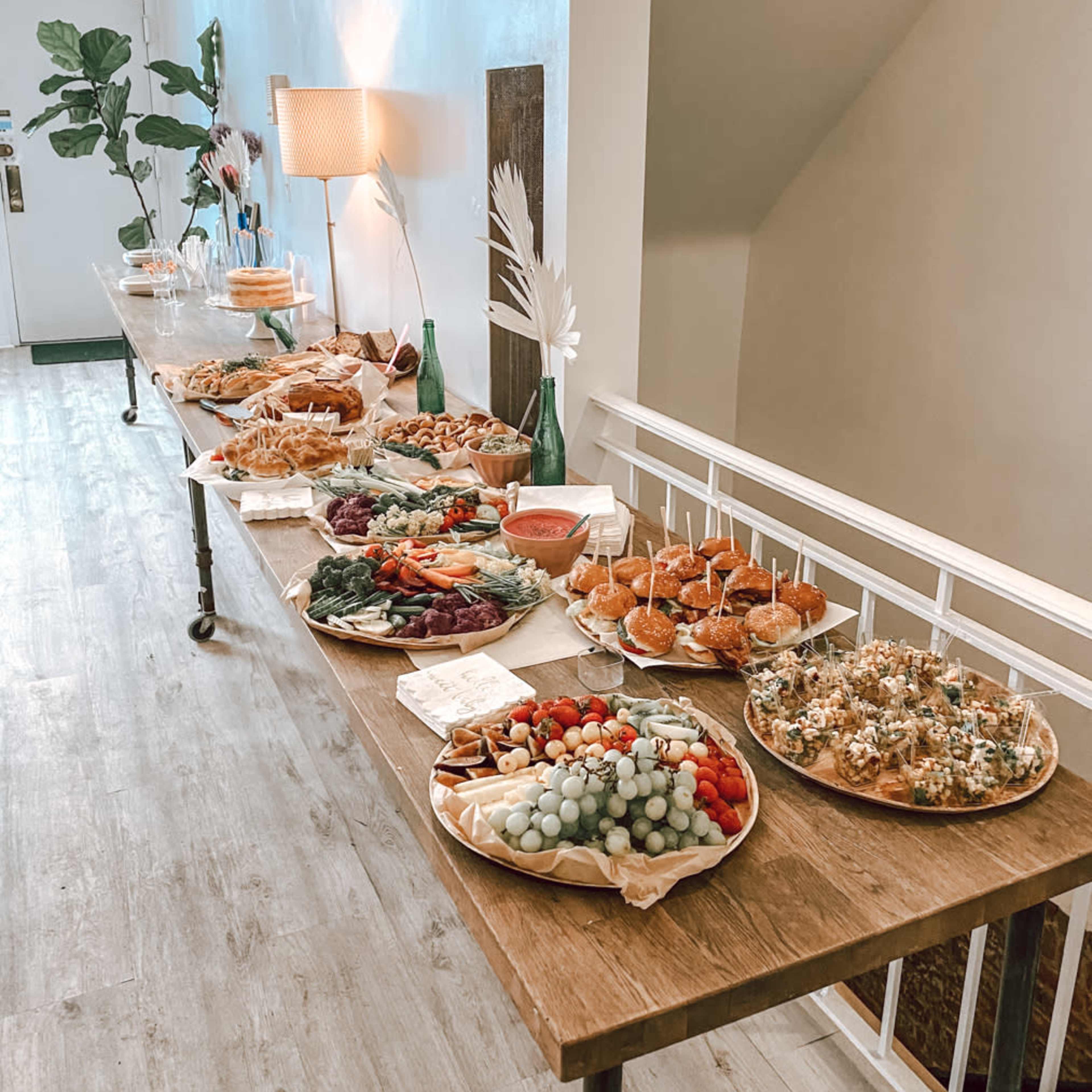 A long wooden table is set with a variety of platters featuring sandwiches, fruits, vegetables, and snacks, arranged in an indoor space.