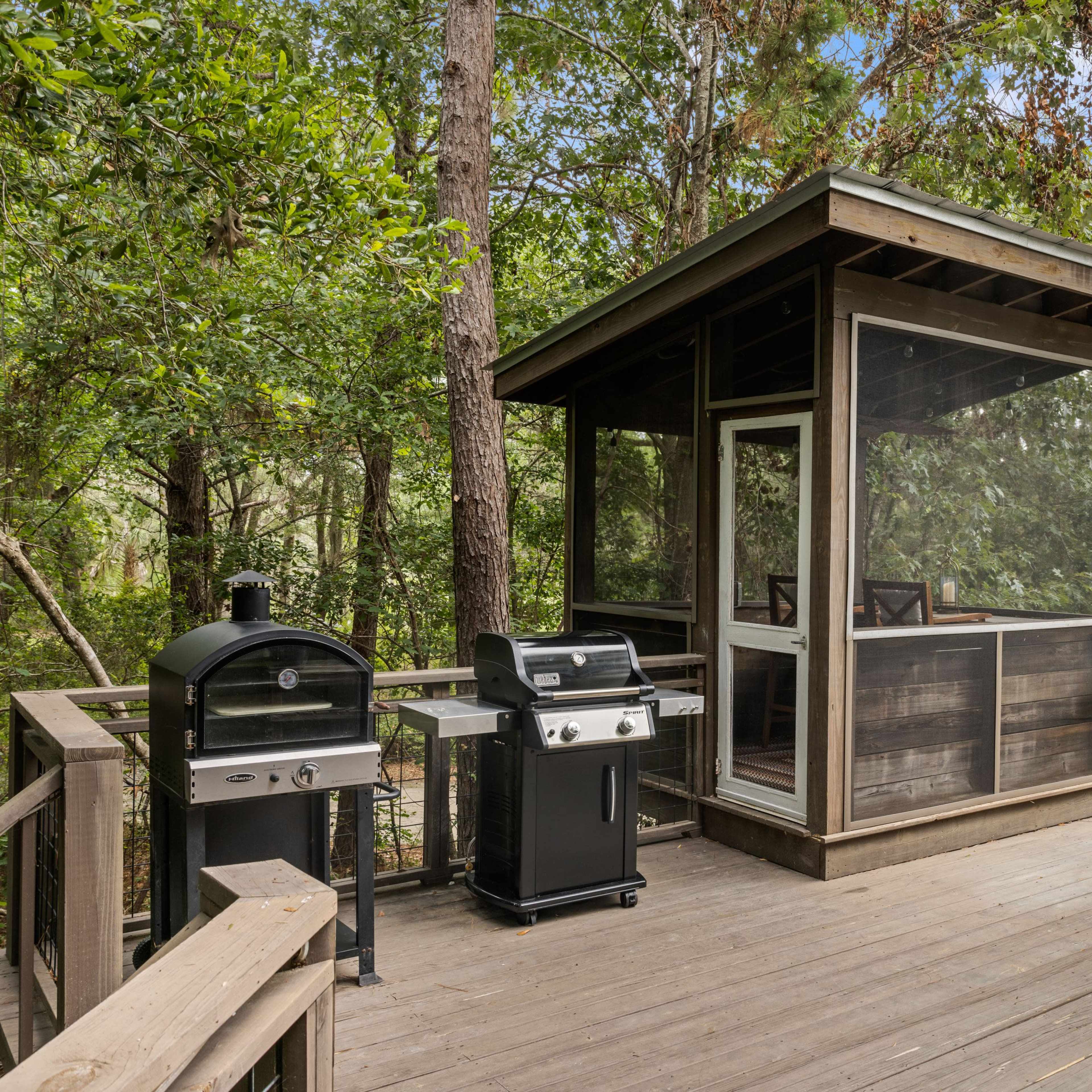 A wooden deck featuring a screened-in gazebo, a charcoal grill, a gas grill, and a hanging chair, all surrounded by trees.