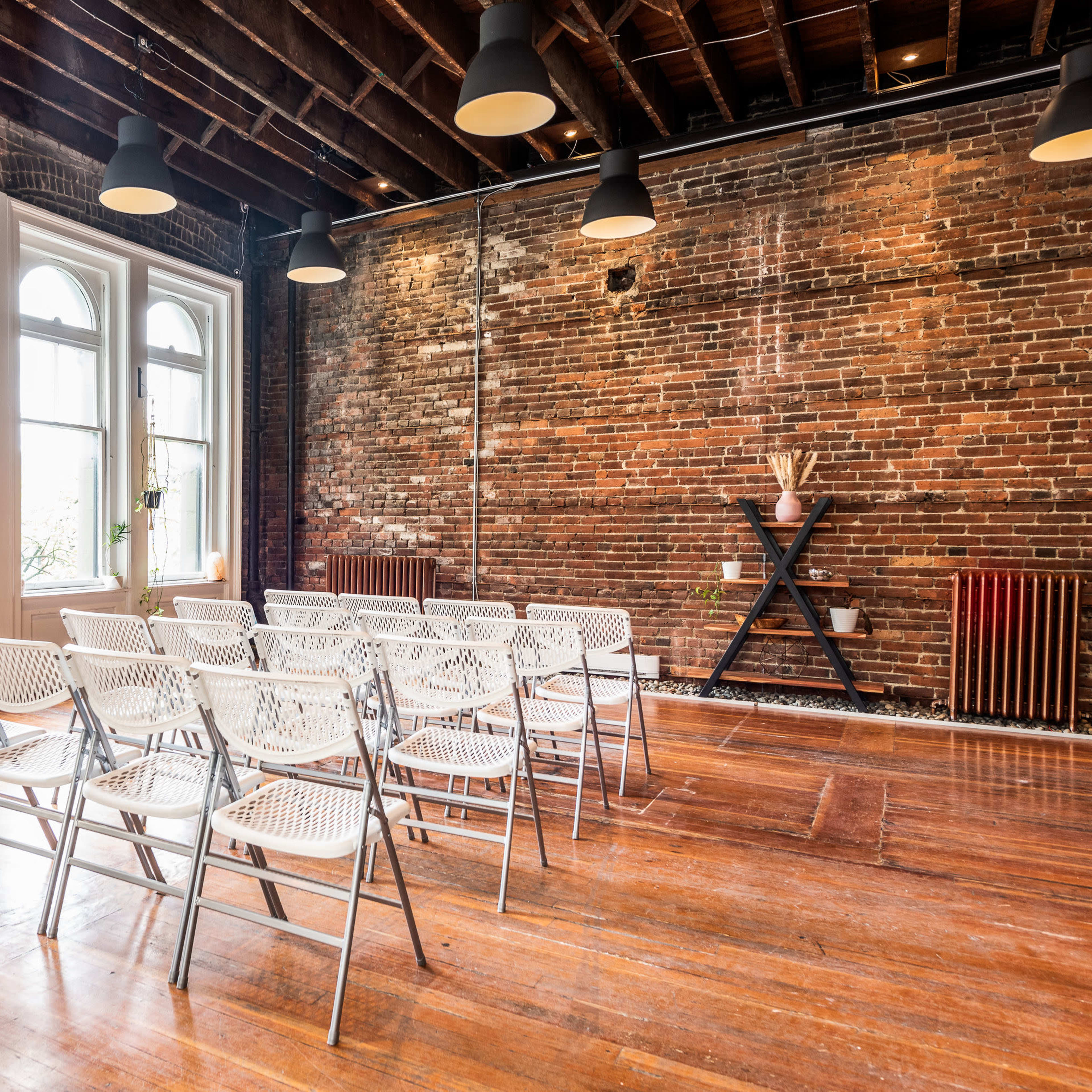 A room with exposed brick walls and wooden floors contains rows of white folding chairs arranged for an event, with large windows and potted plants along the walls.