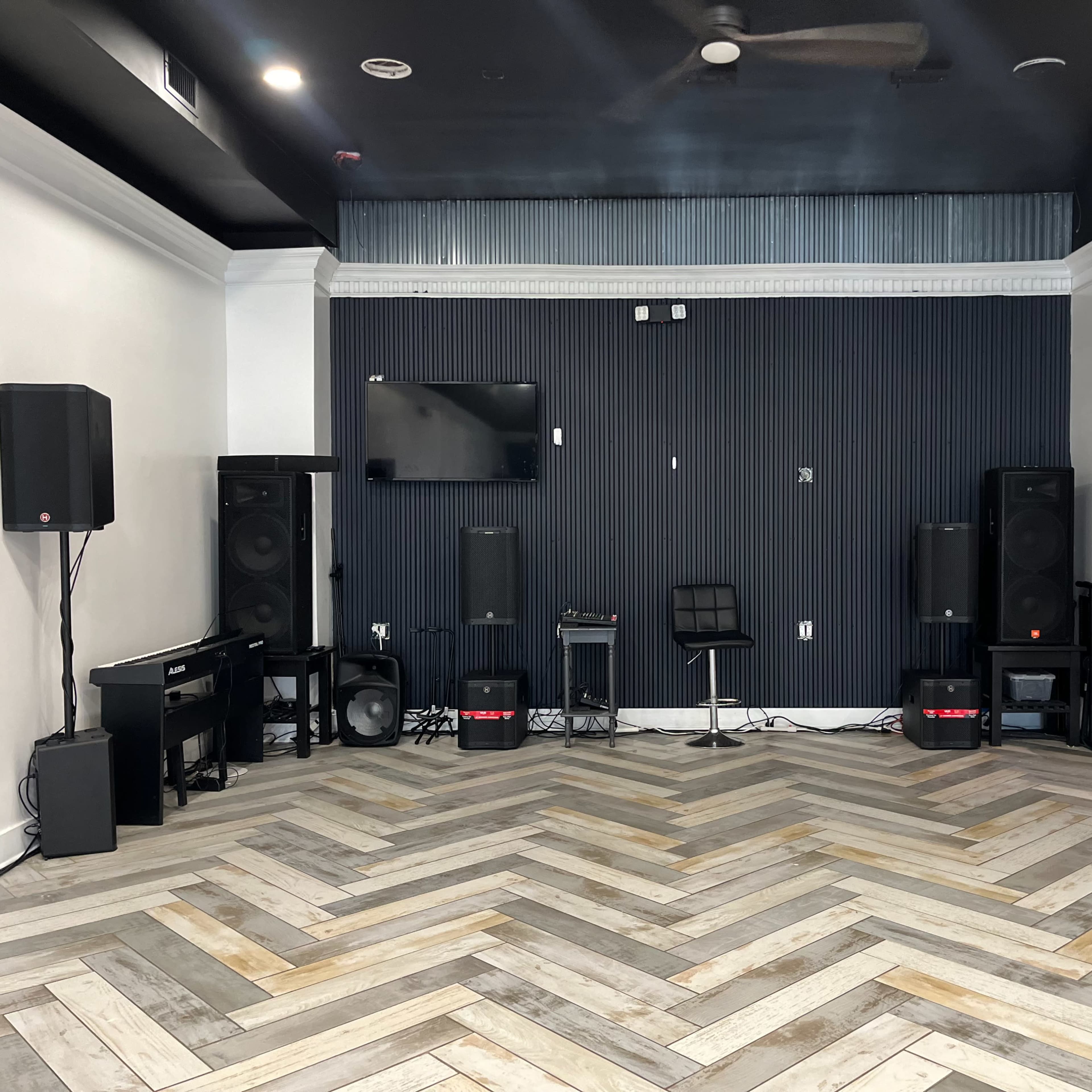 A sparsely furnished room with a herringbone wood floor, featuring speaker stacks, a television mounted on the wall, and a black chair positioned in the center.