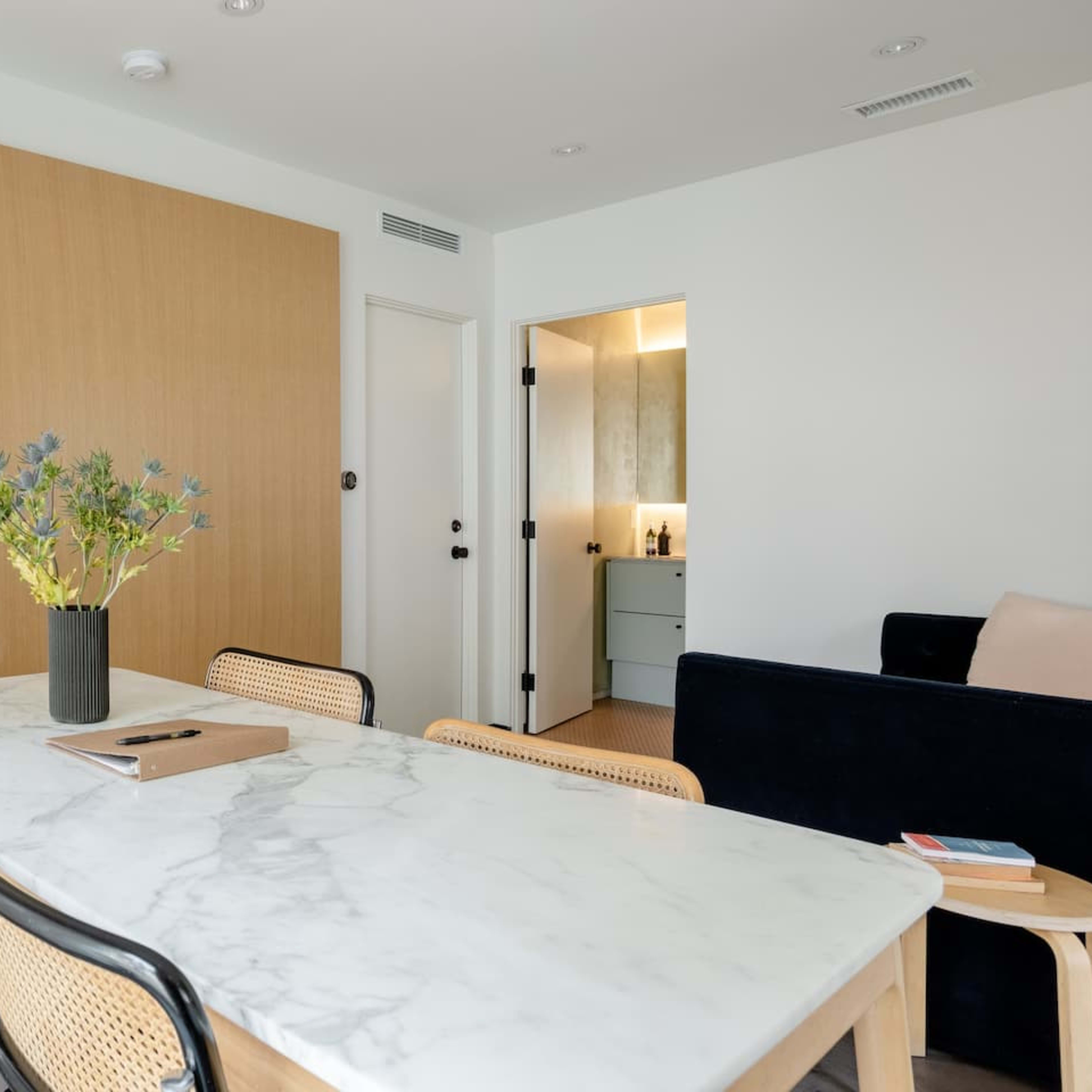 A modern living space features a marble dining table with wooden chairs, a bedroom with a bed visible through a sliding door, and a bathroom entrance in the background.