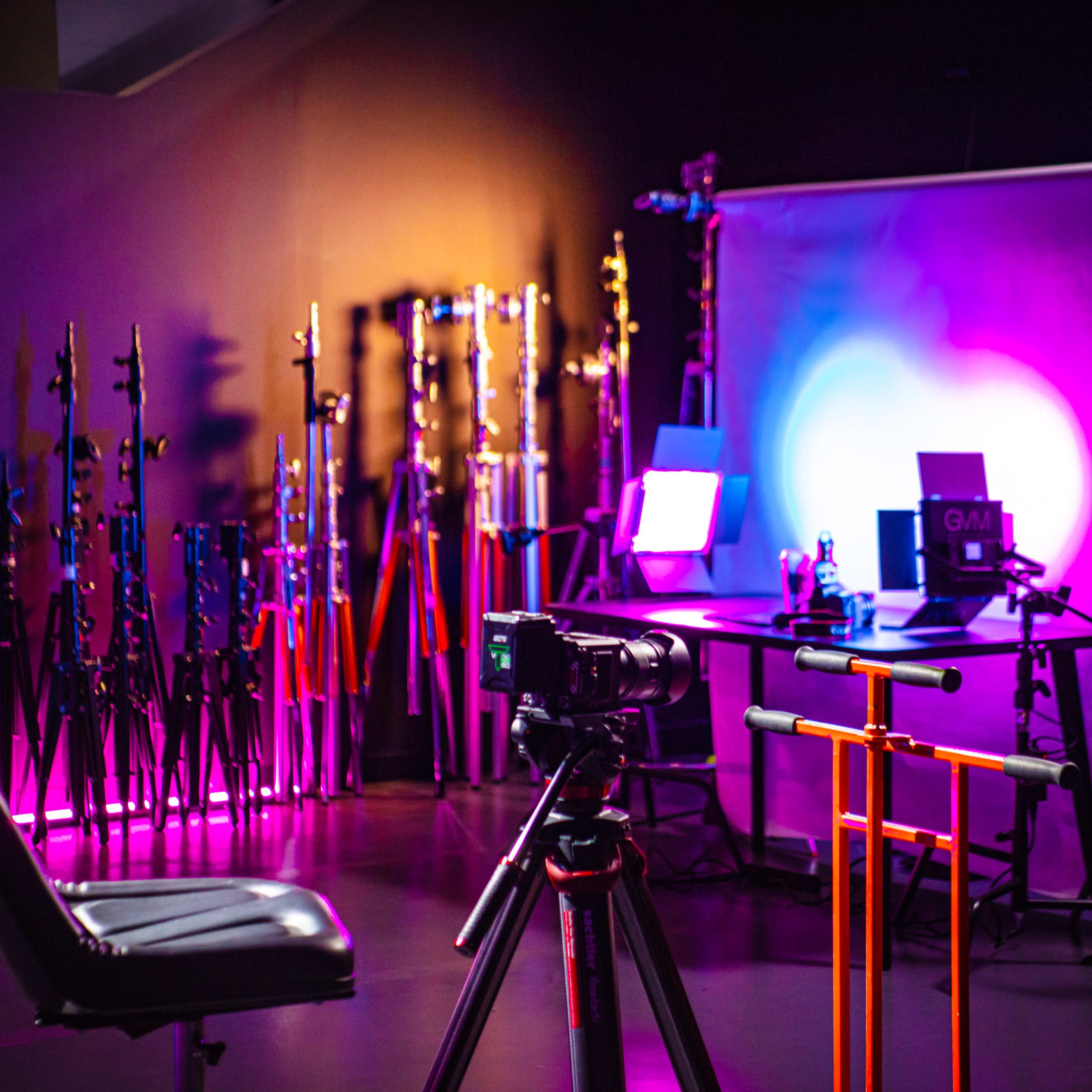 A camera is positioned on a tripod facing a backdrop with lighting equipment and a chair in a studio setting.