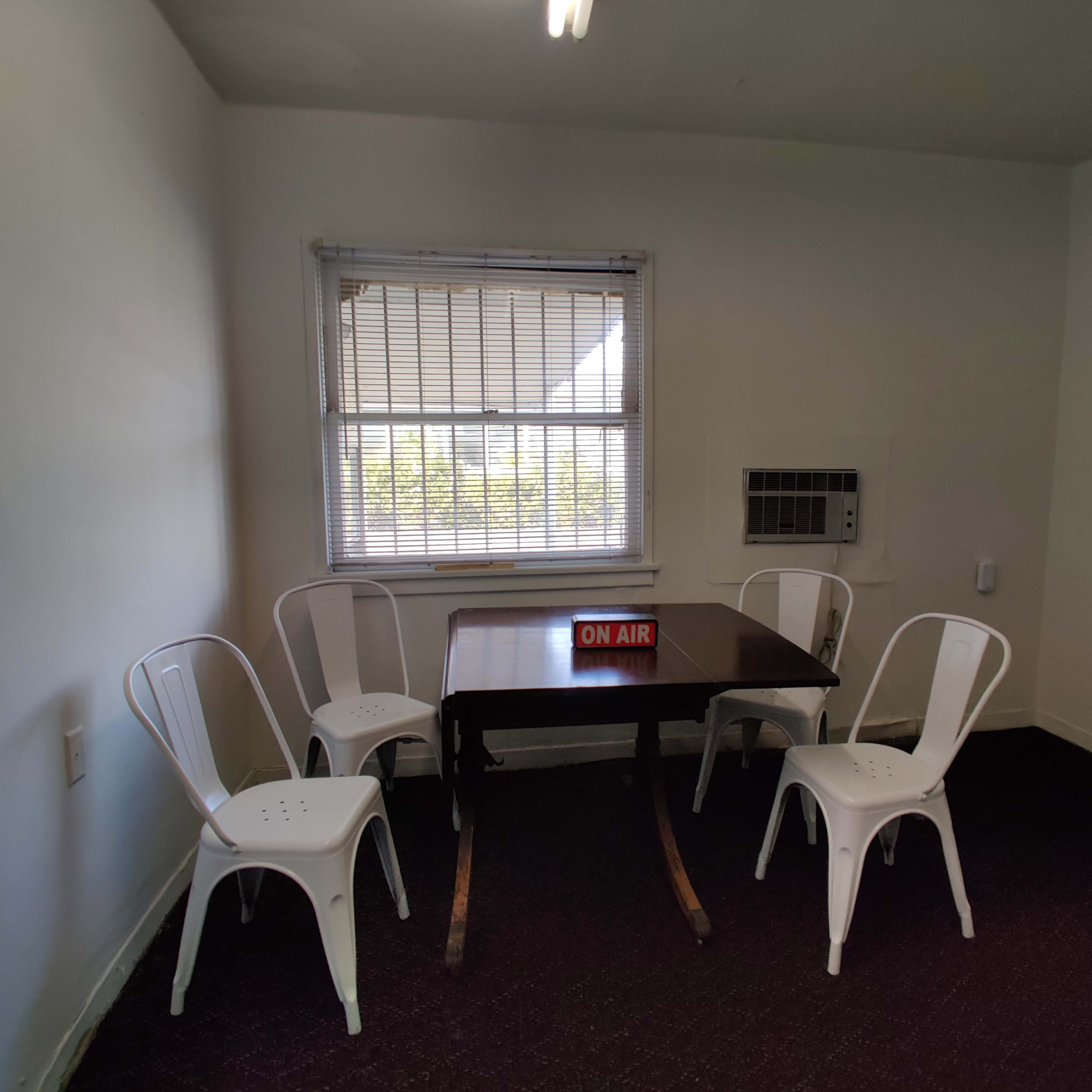A small room features a table with four white chairs, a window with blinds, and an "ON AIR" sign.
