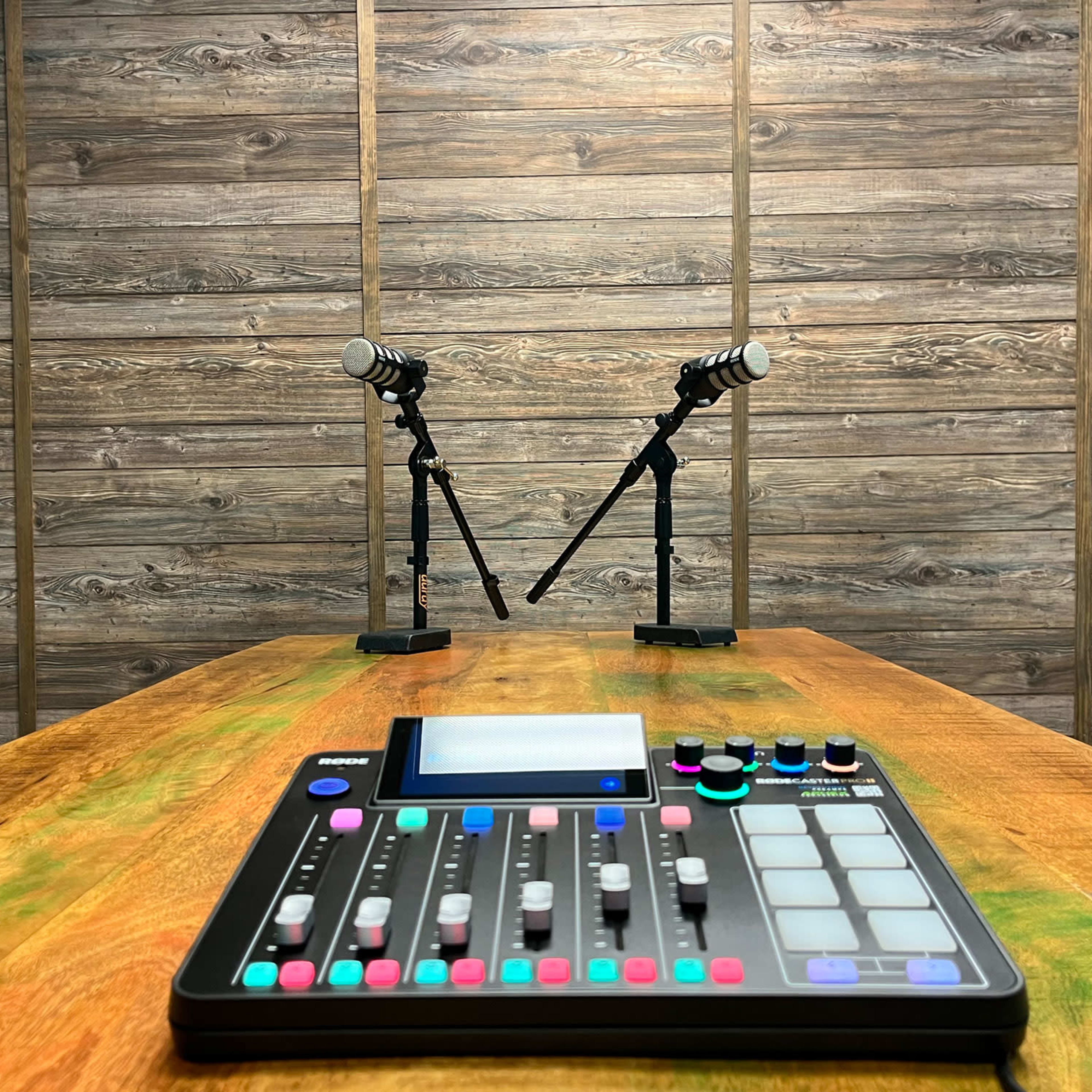 A wooden table with a digital audio mixer in the foreground and two microphones on stands set up against a wooden wall.