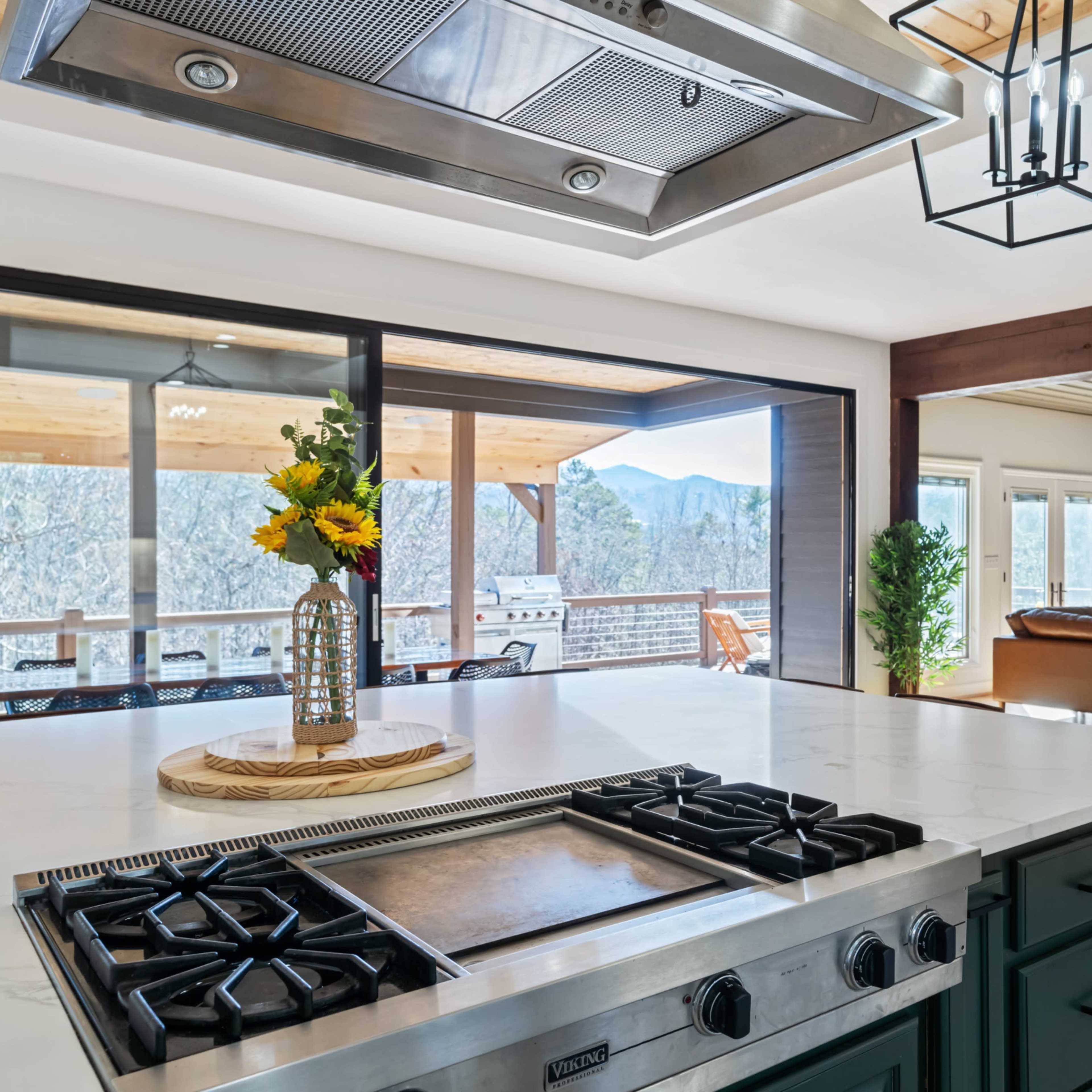 A modern kitchen featuring a gas stove on a marble countertop, overlooking a spacious living area and a porch with mountain views.