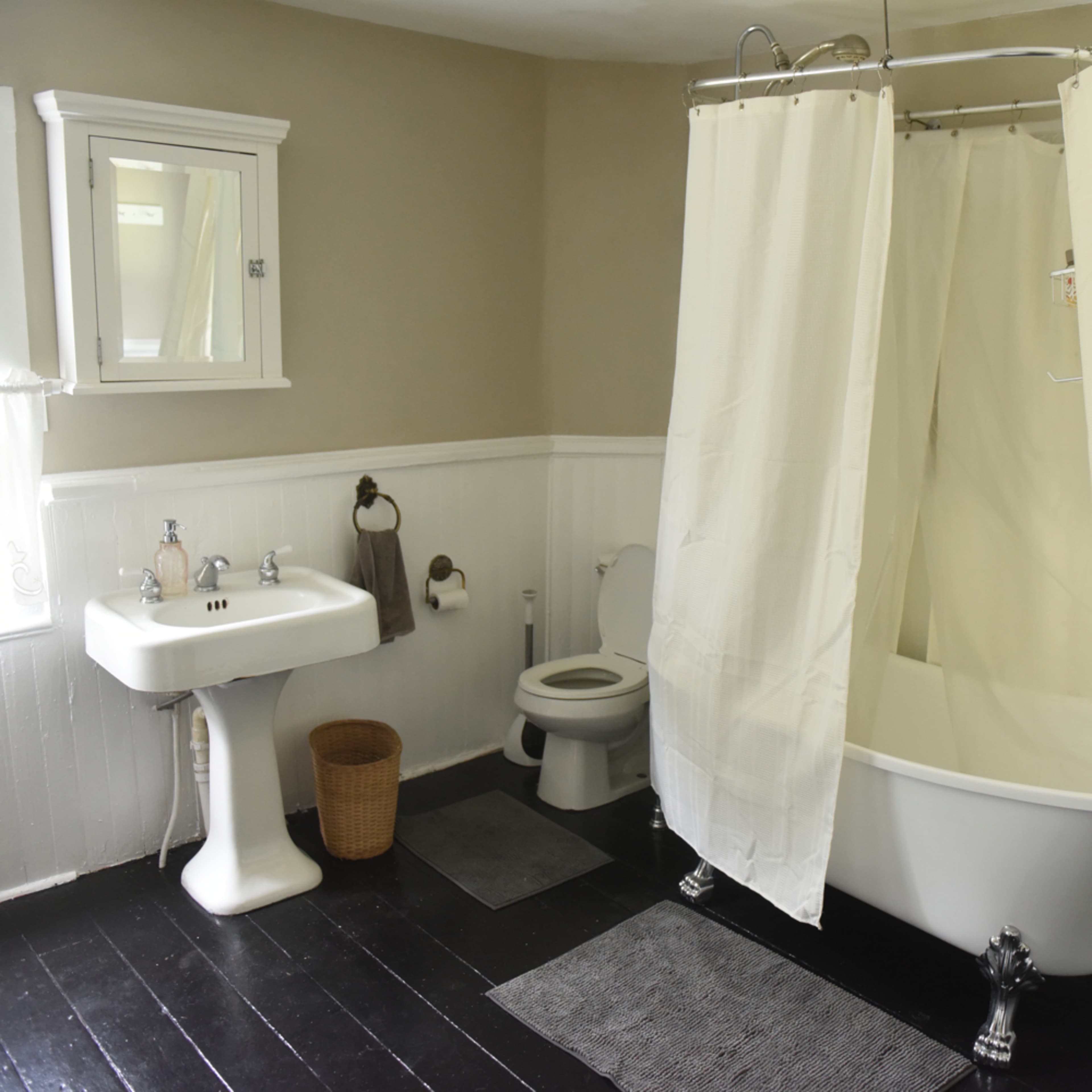 A bathroom featuring a white sink, a toilet, a freestanding bathtub with a shower curtain, and two windows.