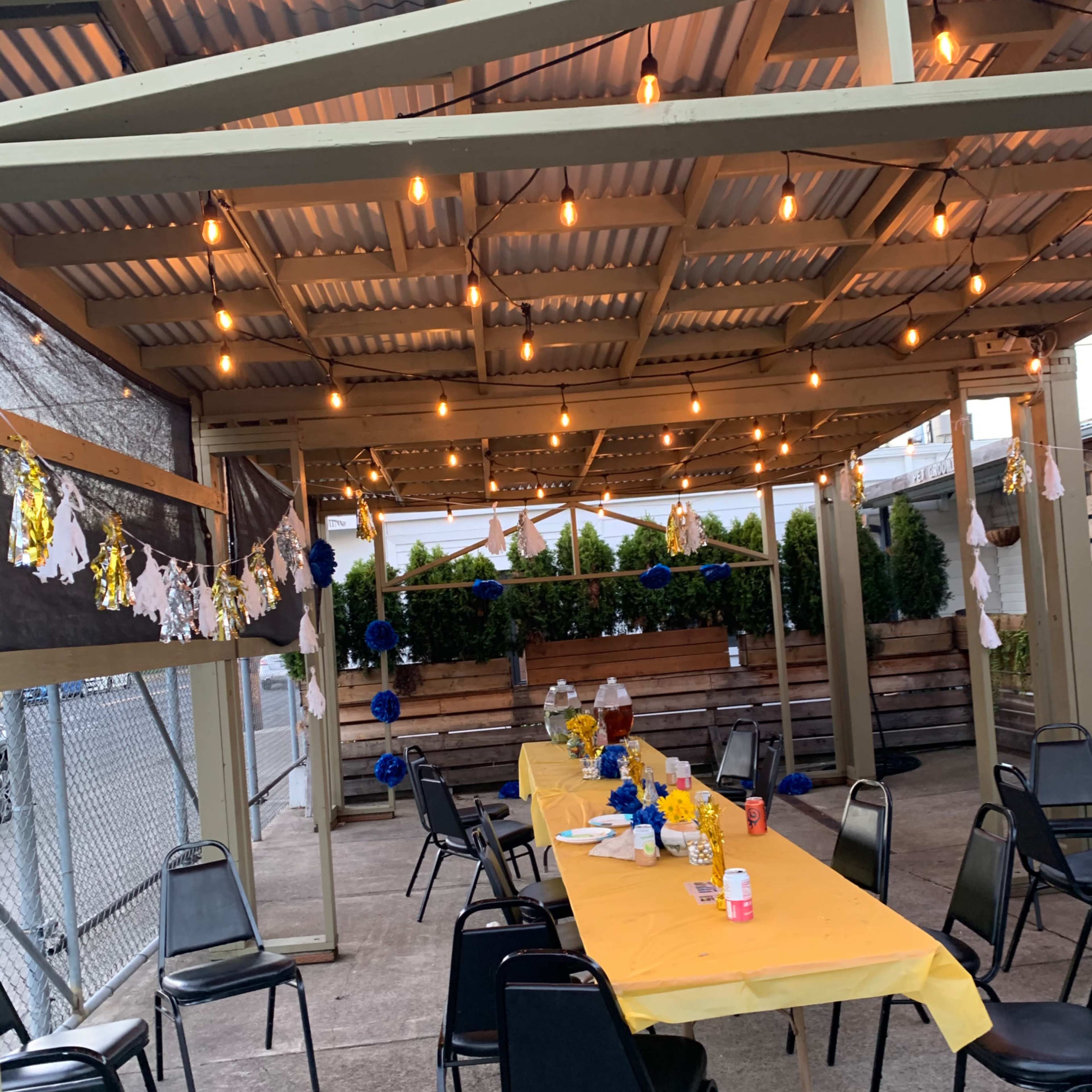 A decorated outdoor space features a long table set with yellow cloth and floral arrangements, under string lights and banners.