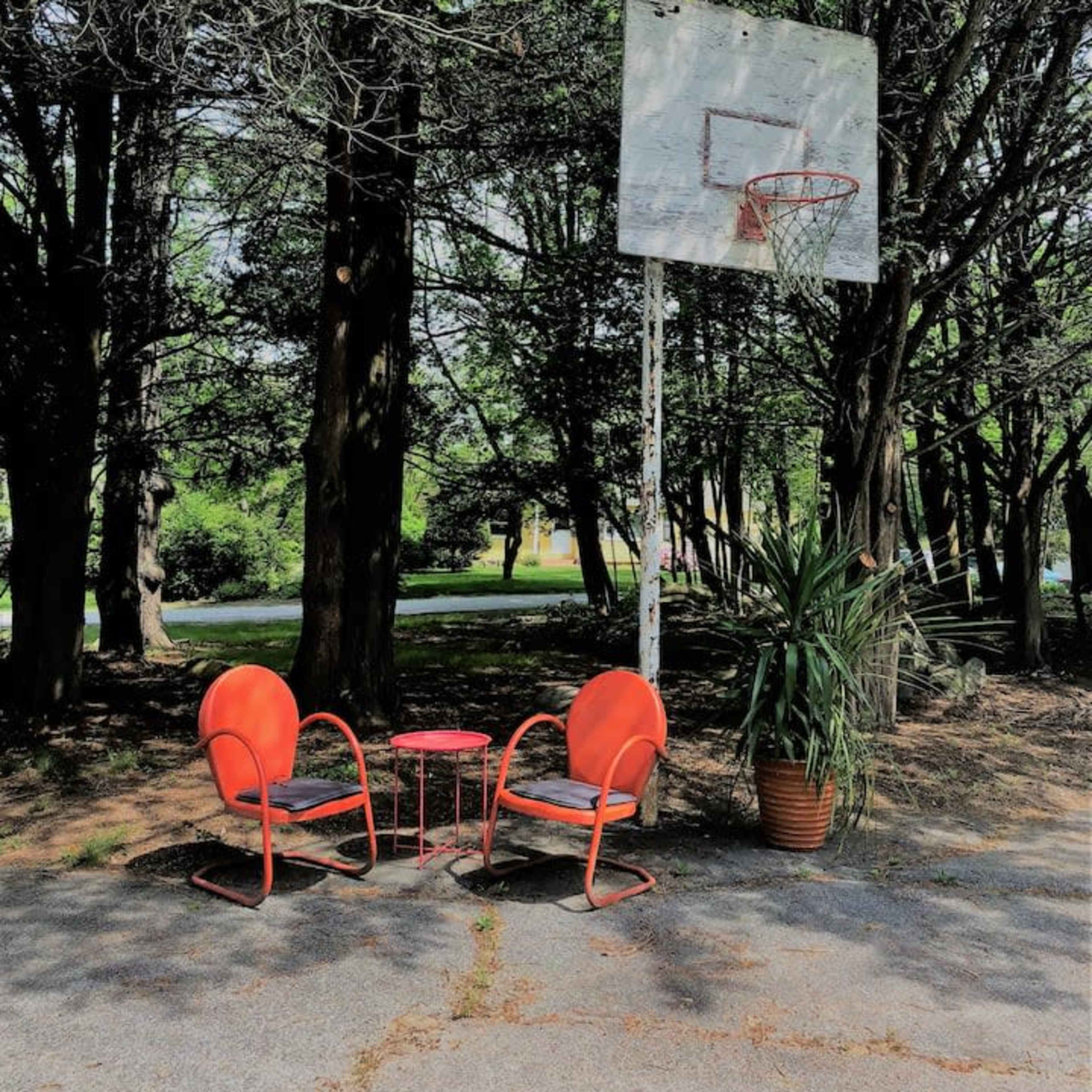 Two orange chairs and a small table are set up near a basketball hoop surrounded by trees.