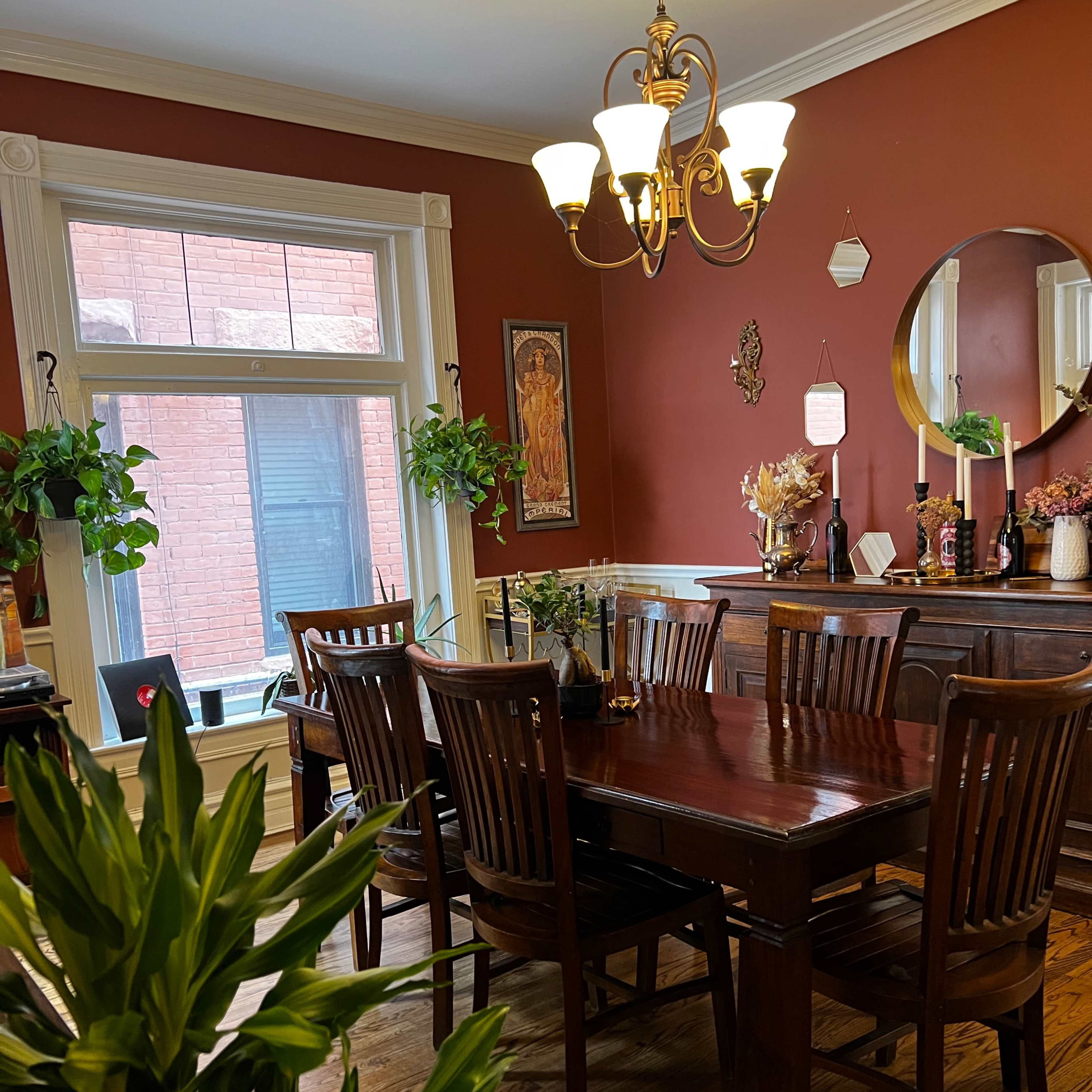 A wooden dining table is surrounded by chairs in a warmly colored room with plants, a chandelier, and a decorative mirror.