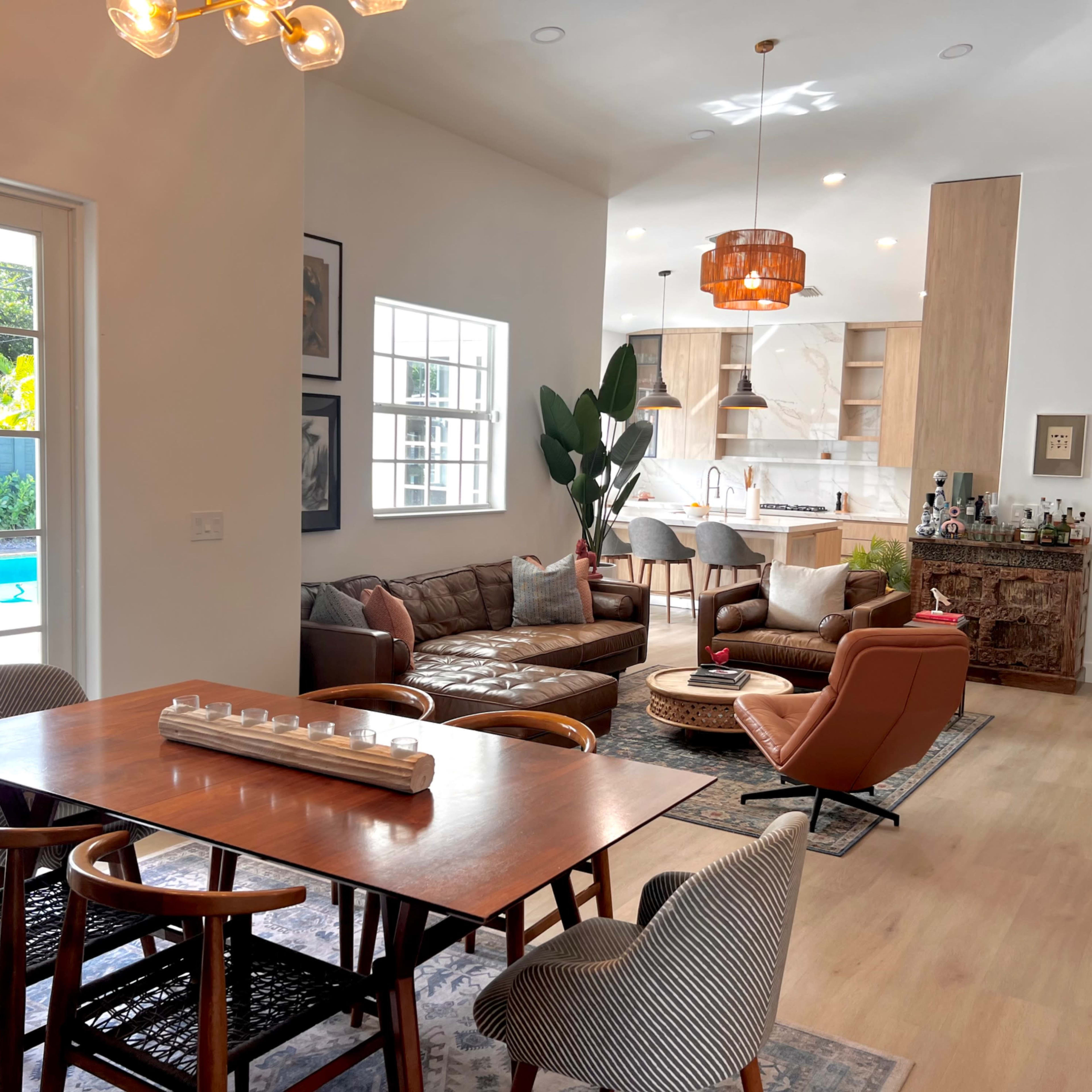 A modern living and dining area with a pool view, featuring a wooden dining table, a brown leather sofa, and a contemporary kitchen in the background.