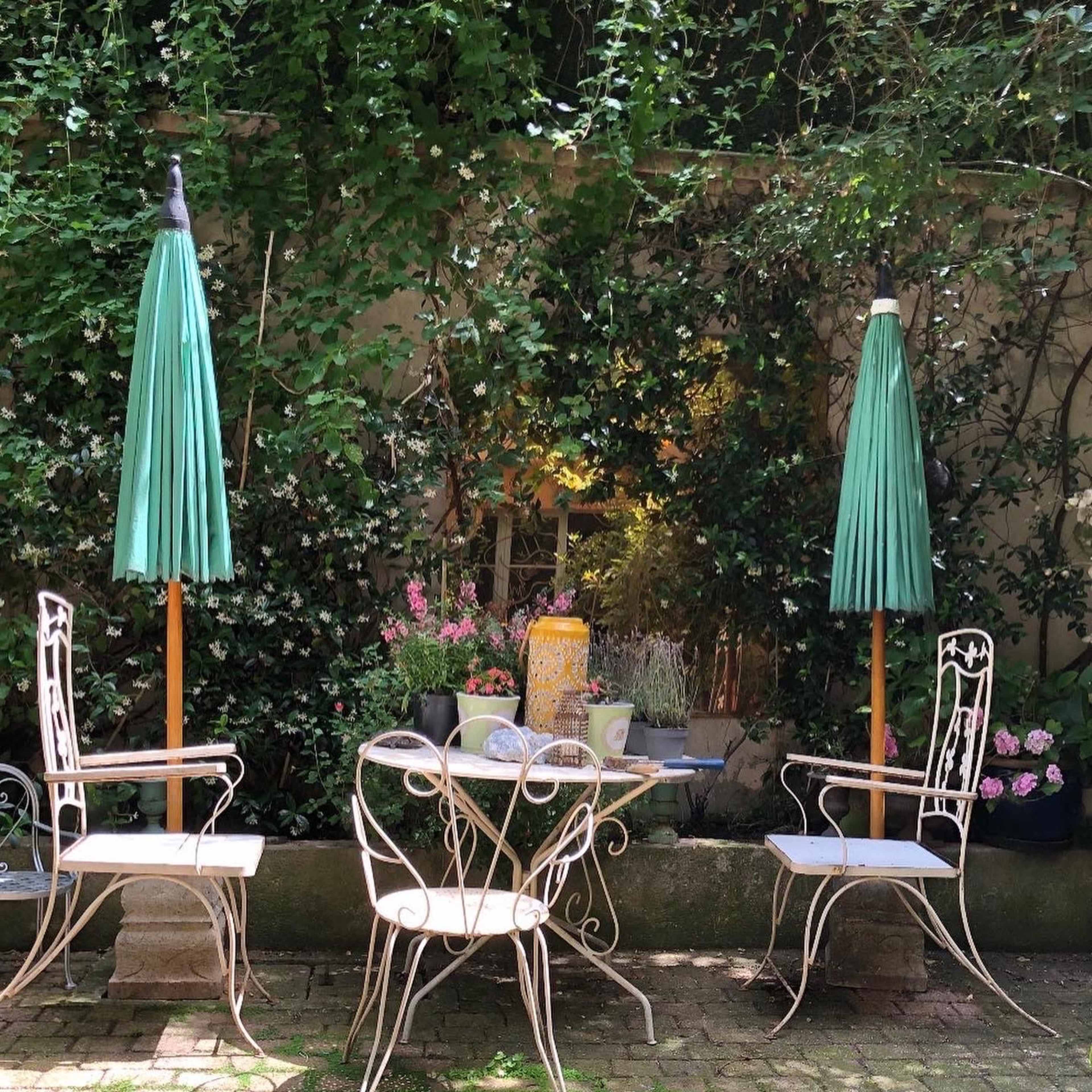A small garden table with two chairs featuring green umbrellas is surrounded by lush greenery and colorful flowers.