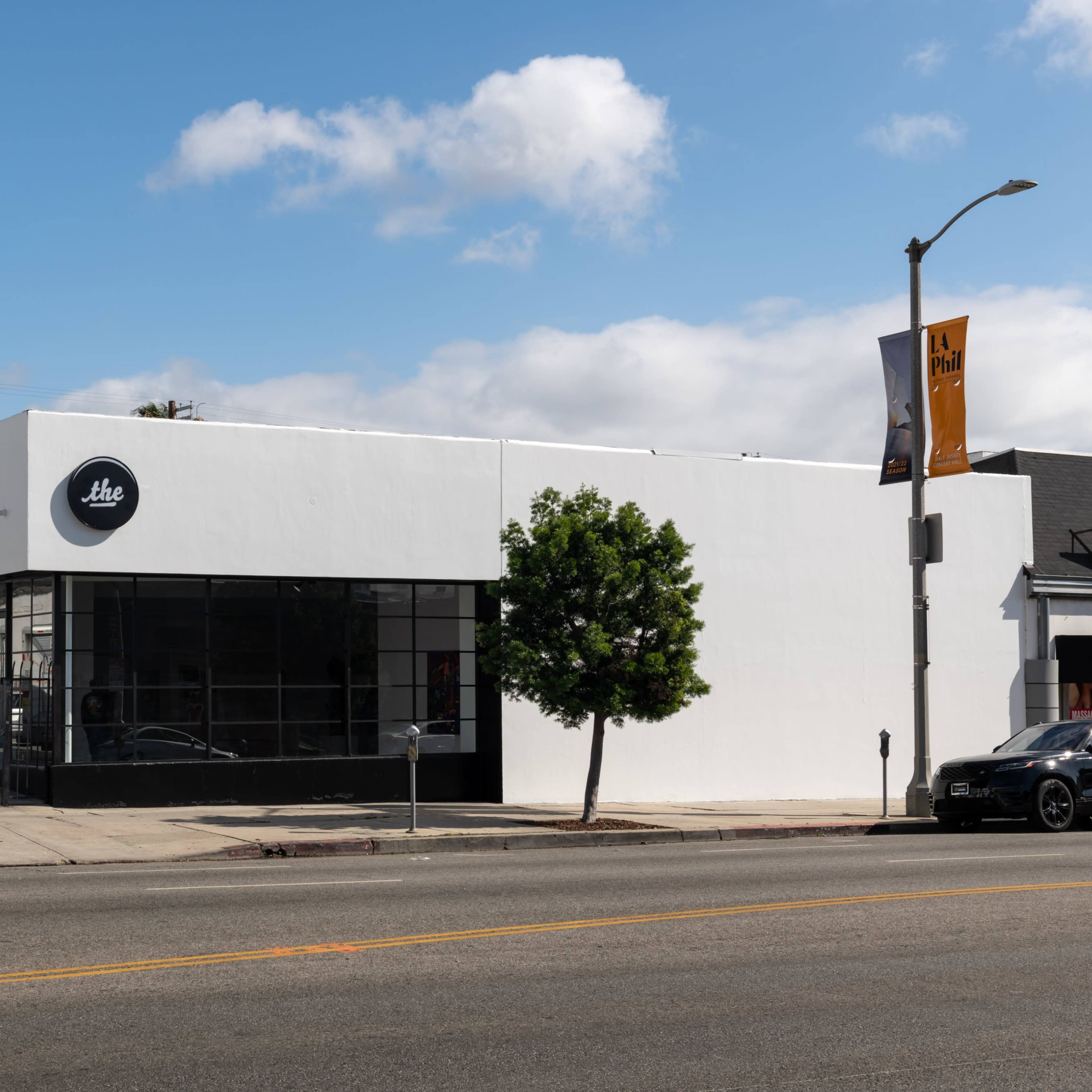 A modern white building with large windows and a circular logo, situated on a street next to a parked car.