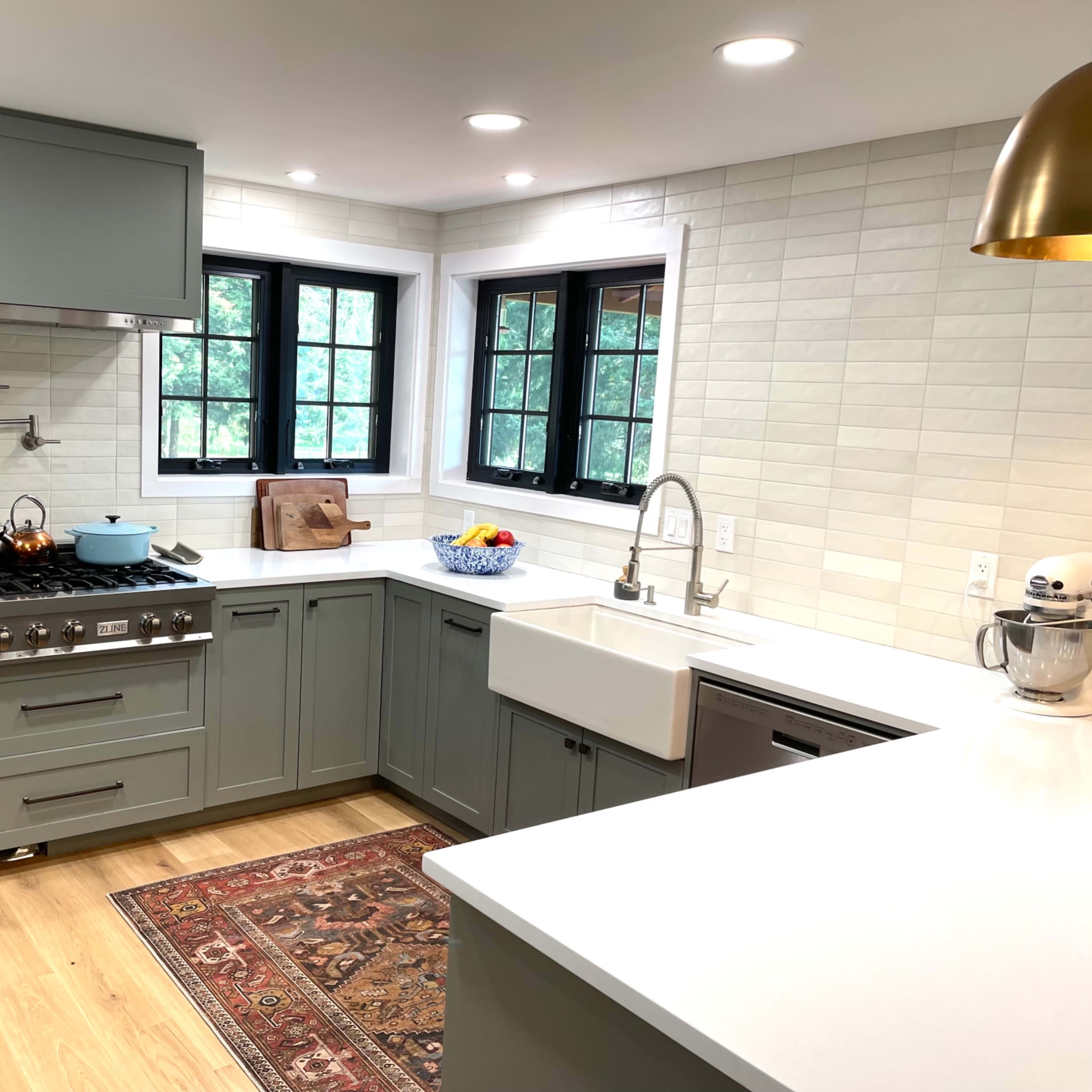 A modern kitchen features gray cabinetry, a large farmhouse sink, stainless steel appliances, and a light-colored countertop with a decorative rug.