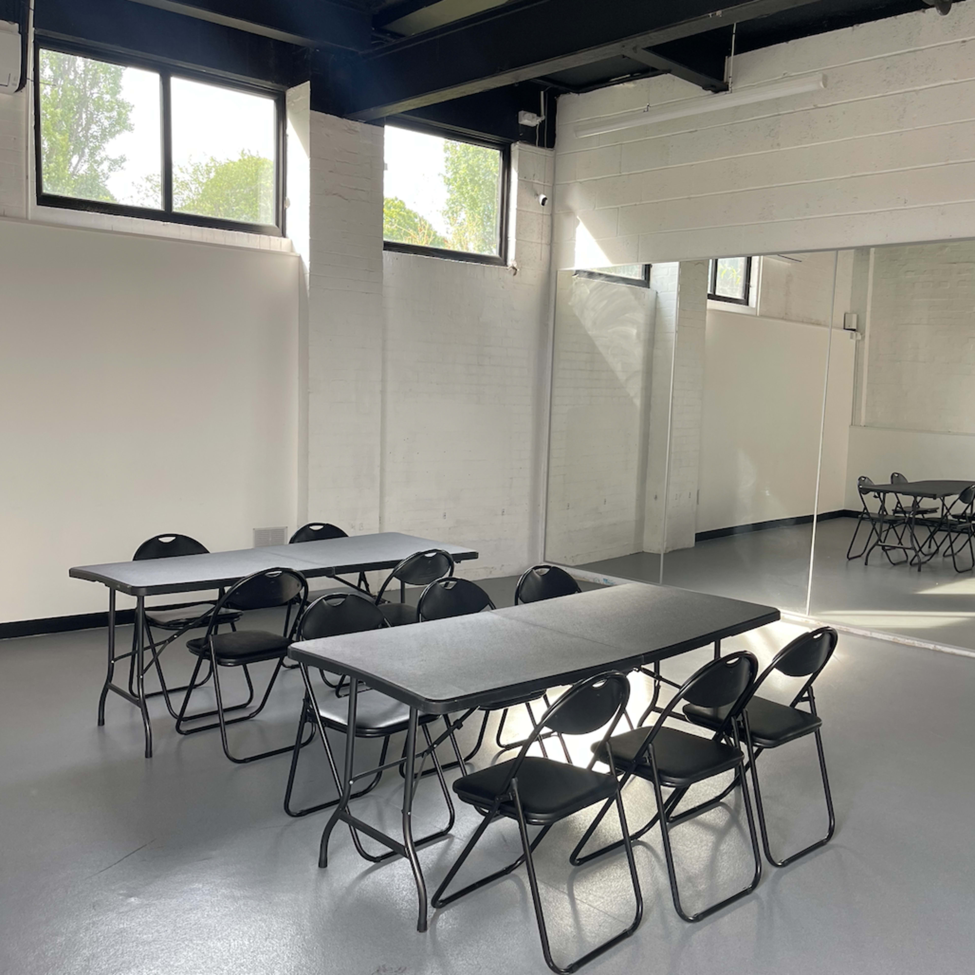 A room with gray flooring contains several folding tables and chairs arranged in a simple layout, with large windows allowing natural light to enter.