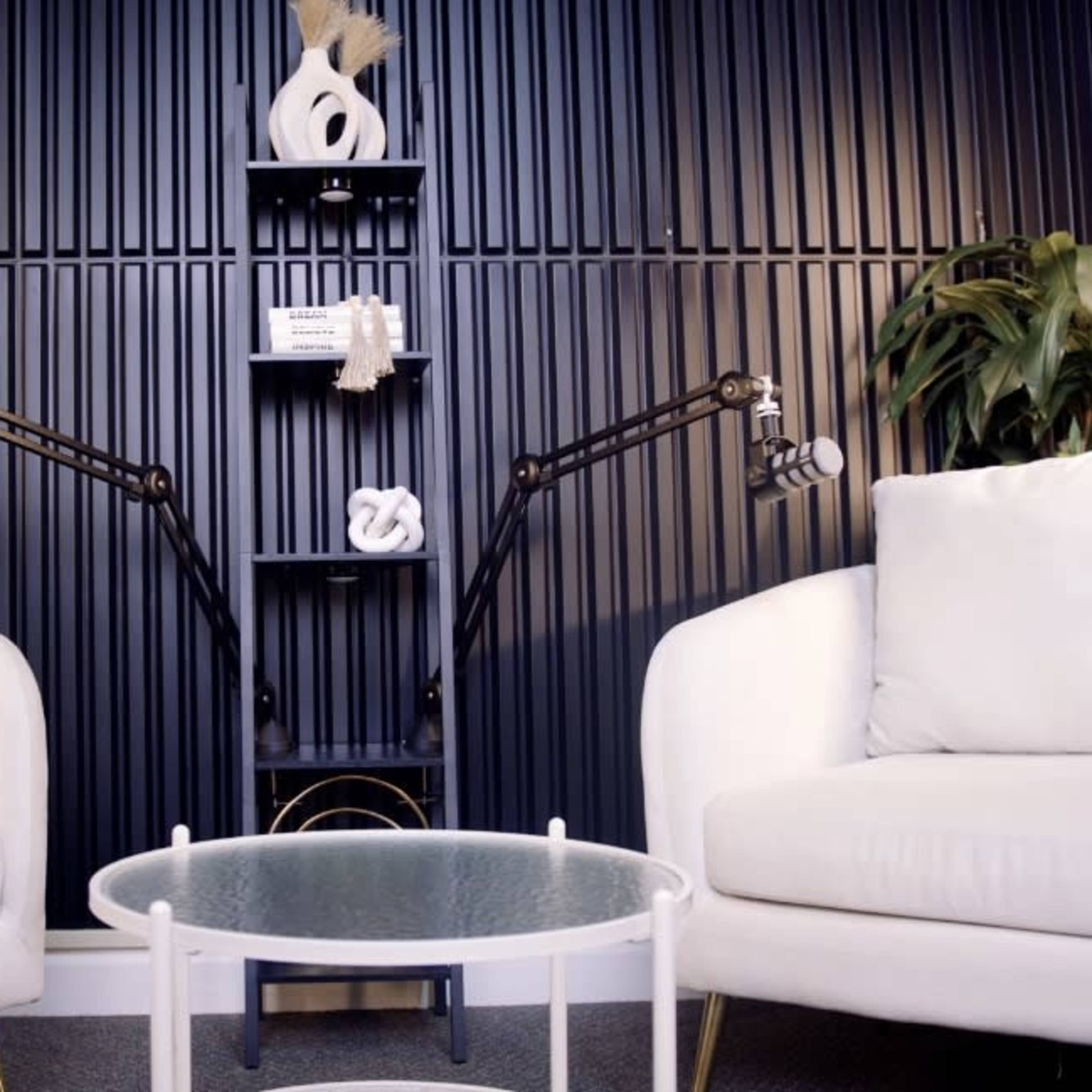 Two white armchairs facing each other with a glass table in between, set against a dark, textured backdrop featuring plants and a modern shelf.