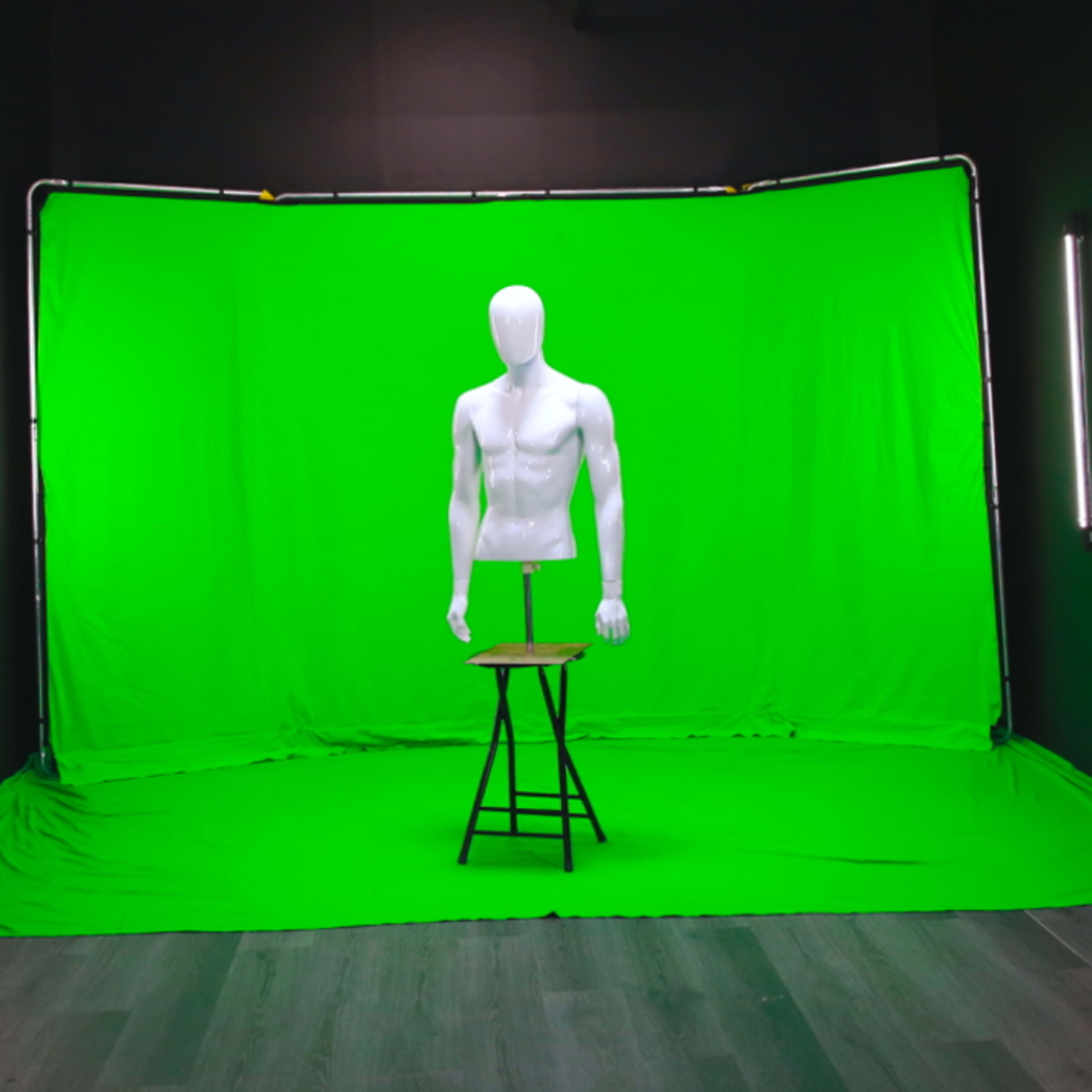 A white mannequin stands on a stool in front of a green backdrop in a dimly lit studio.
