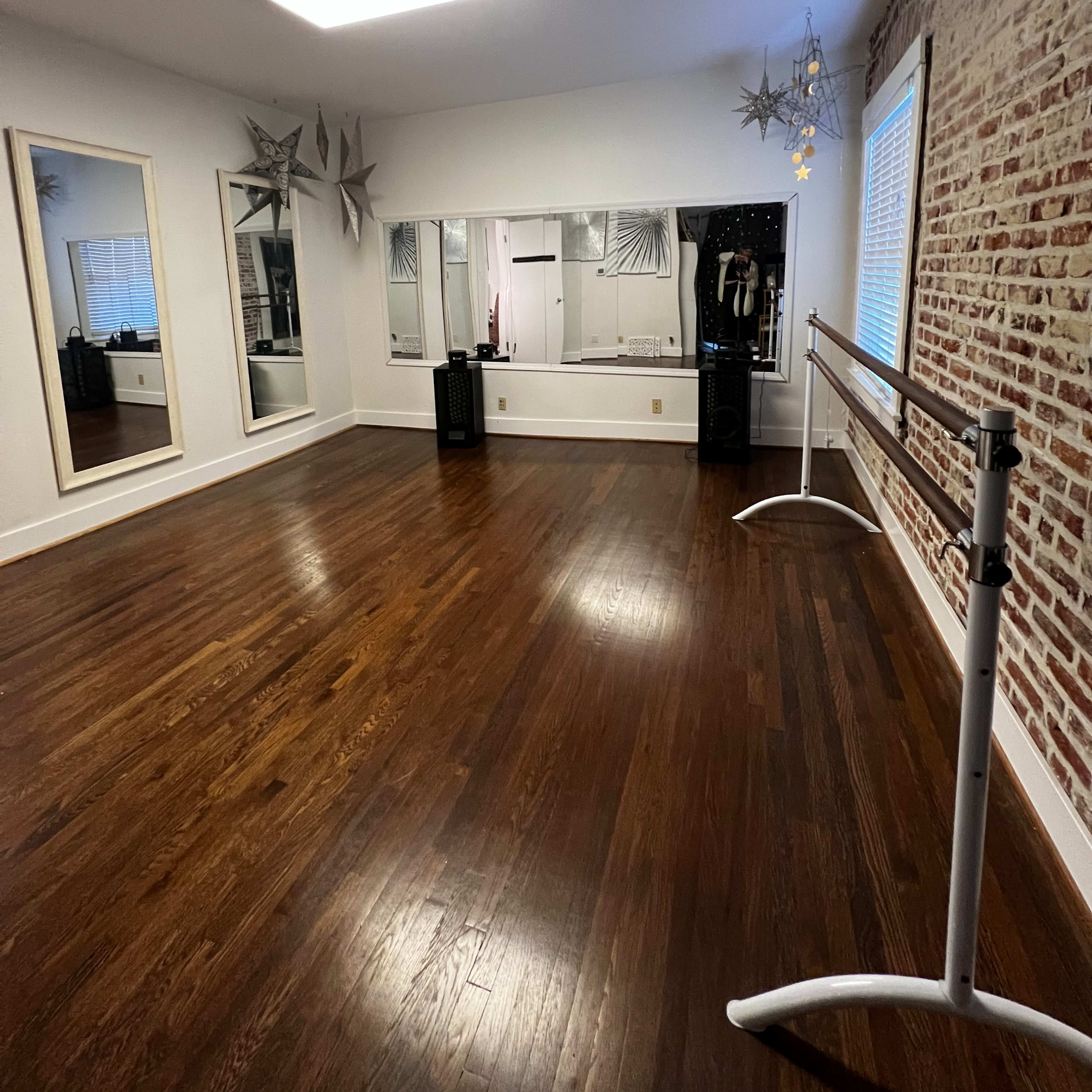 A dance studio with wooden flooring, decorative mirrors, a ballet barre, and a large illuminated ceiling fixture.