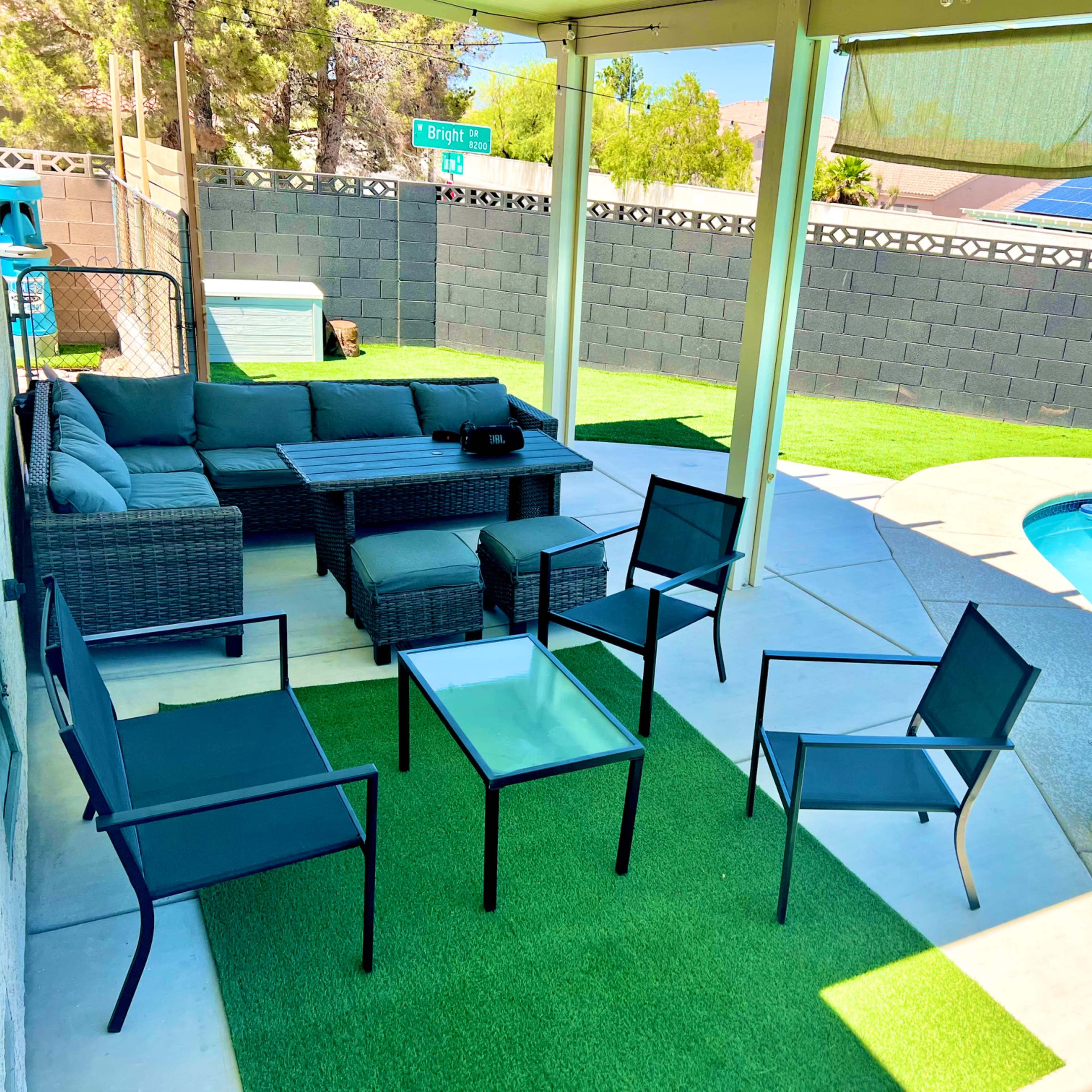 A patio area features a seating arrangement with black chairs and a table, overlooking a pool surrounded by artificial grass and a stone wall.