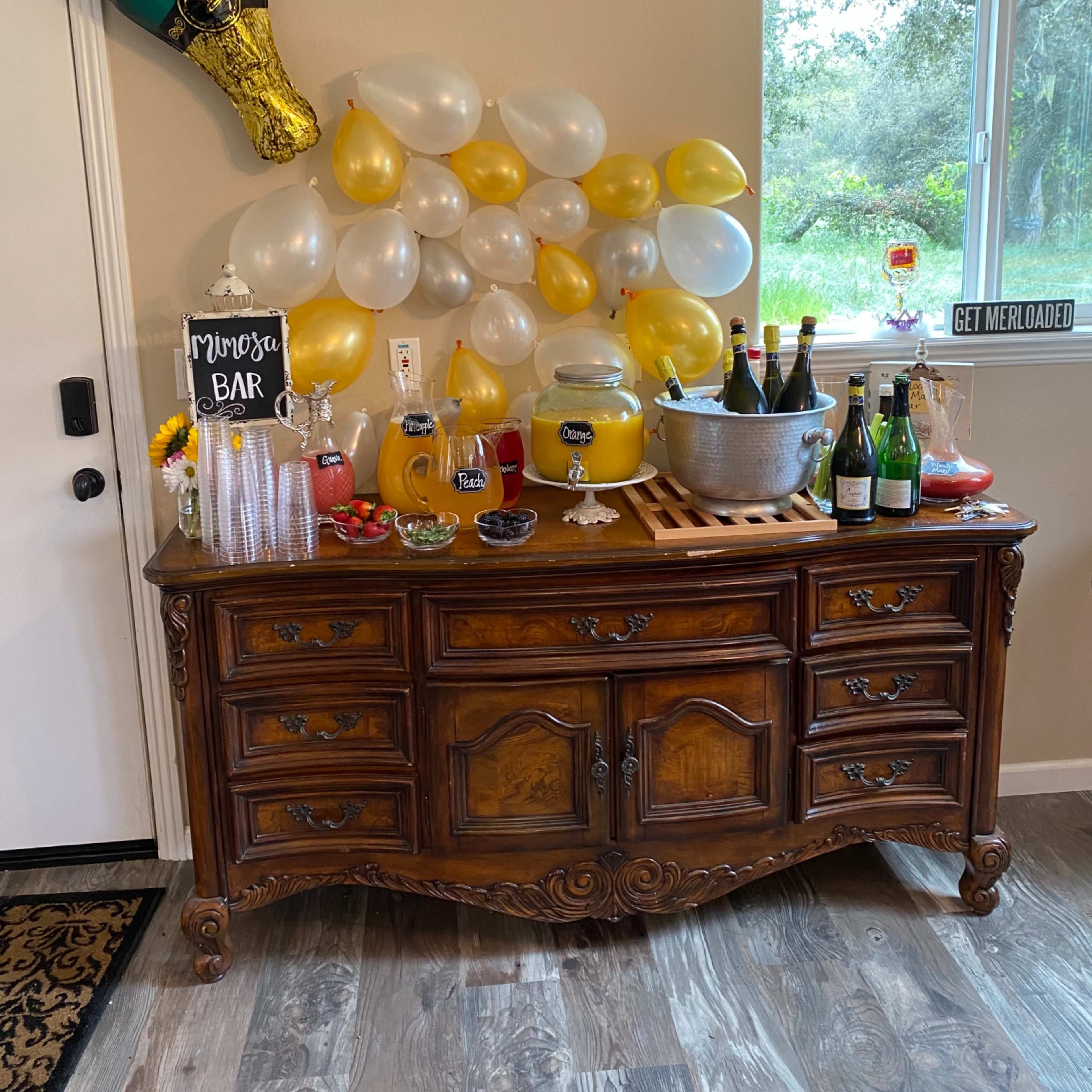 A wooden sideboard displays a variety of drinks, a decorative sign for a mimosa bar, and a backdrop of yellow and white balloons.