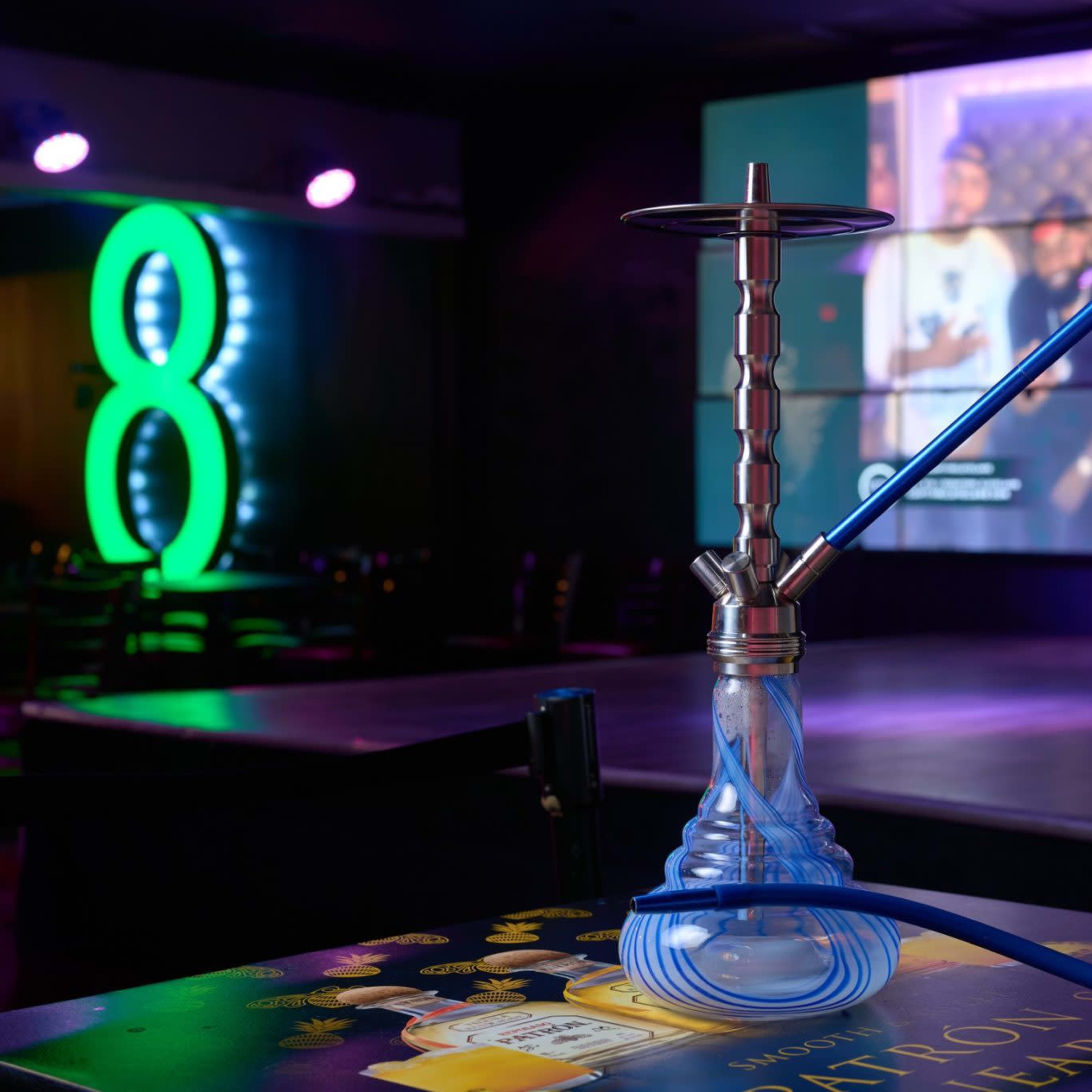 A hookah with a blue hose is placed on a table in a dimly lit venue featuring a large screen in the background displaying a video.