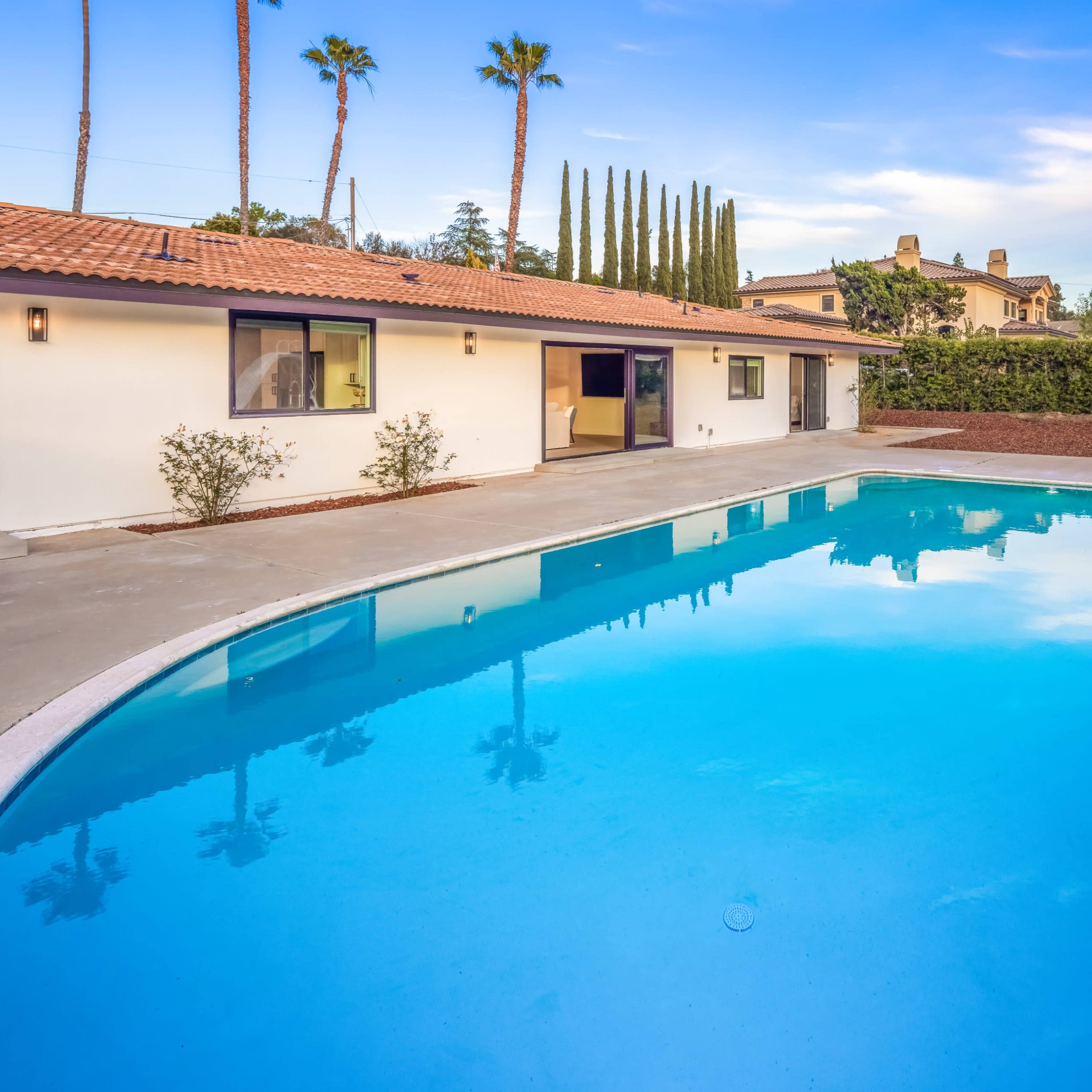 A residential backyard featuring a swimming pool with a slide and a single-story house surrounded by palm trees and manicured landscaping.