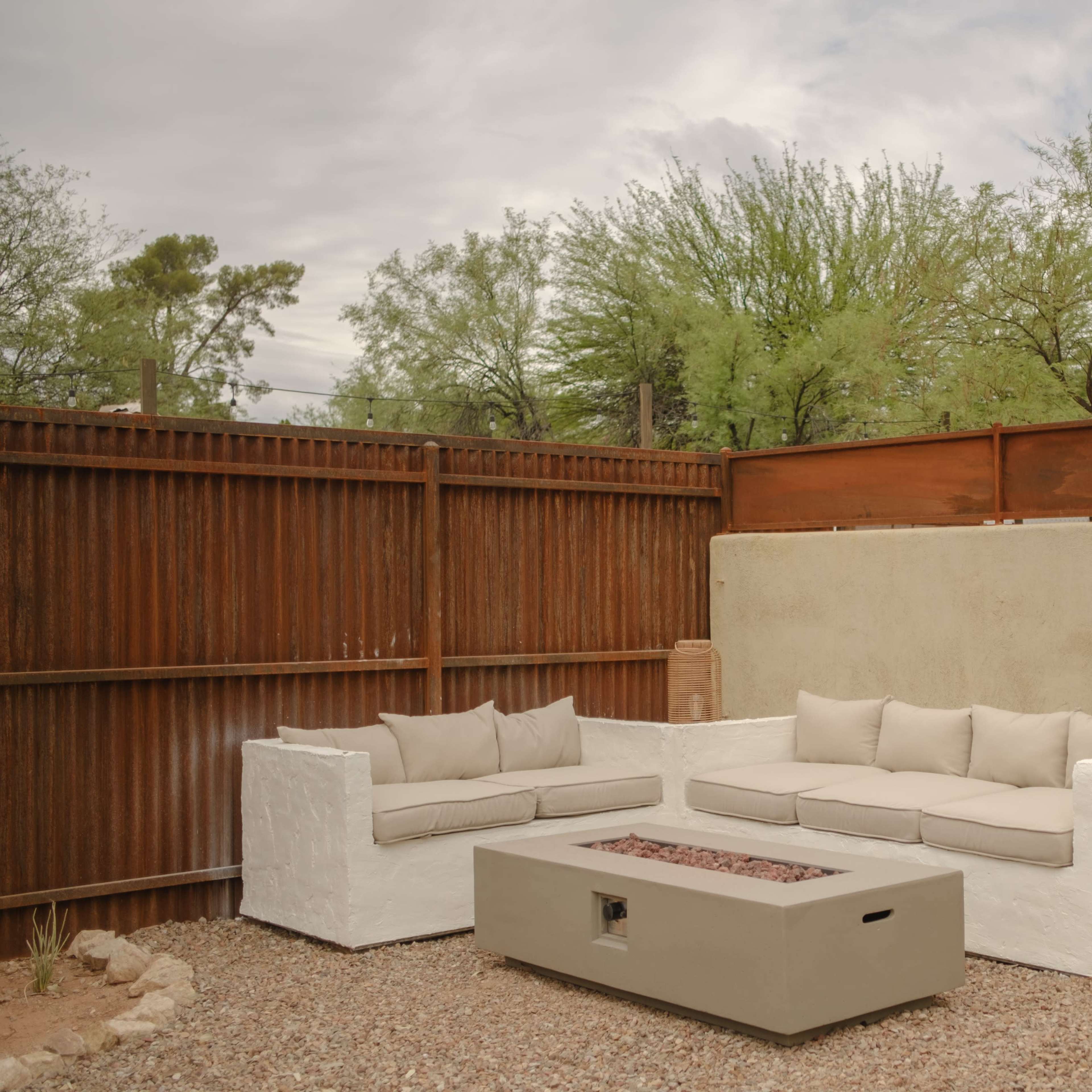 A modern outdoor seating area features a white sectional sofa with beige cushions and a matching rectangular fire pit, surrounded by gravel and greenery against a backdrop of a metal fence.