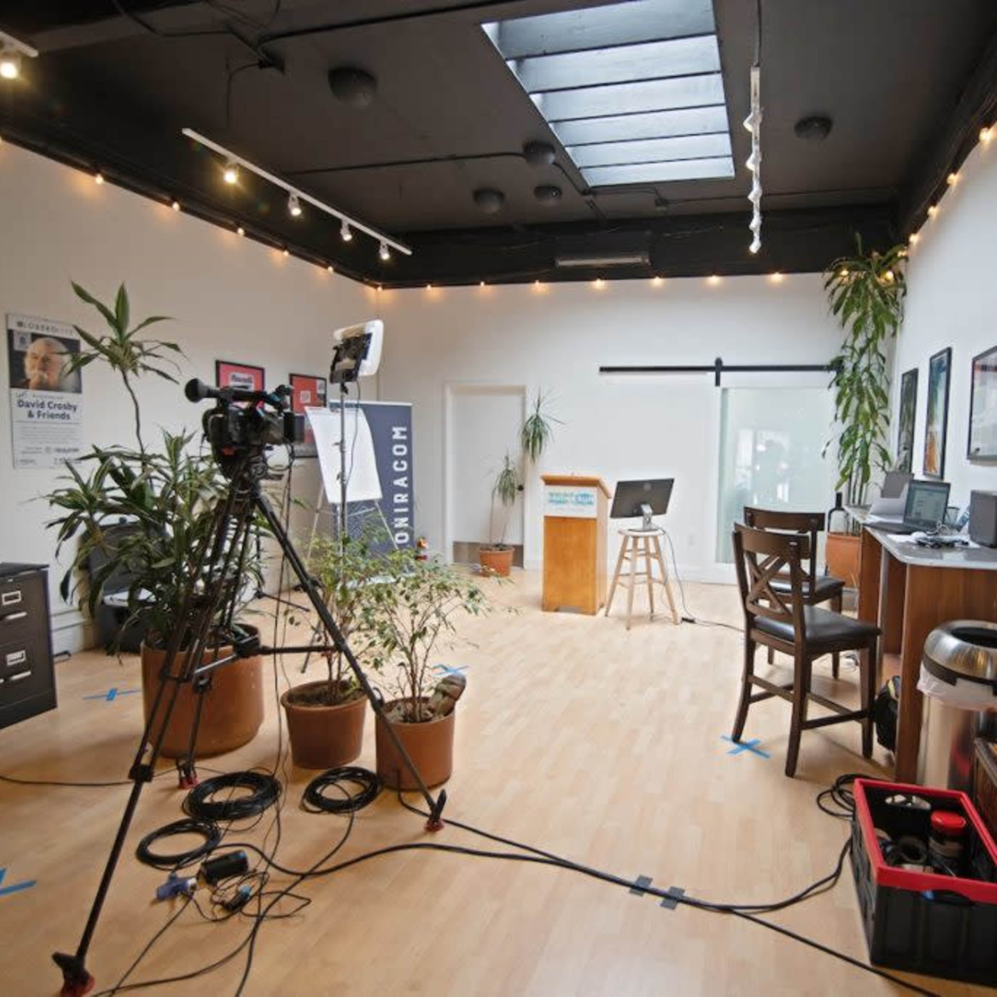 A well-lit studio space with a camera setup, a wooden stool, a desk with a computer, and potted plants throughout.