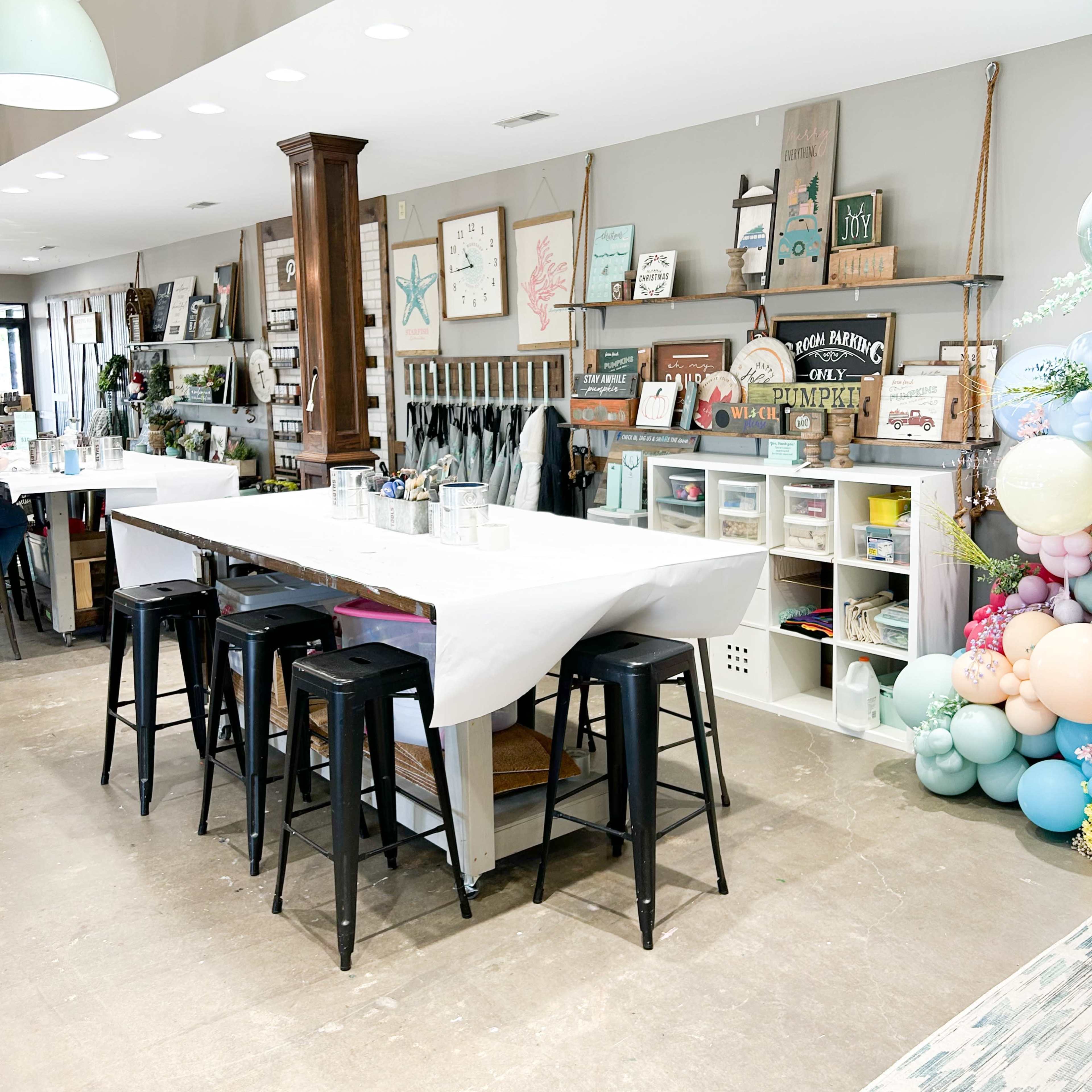 A spacious room features a long table with stools, surrounded by shelves displaying various decorative items and artwork.