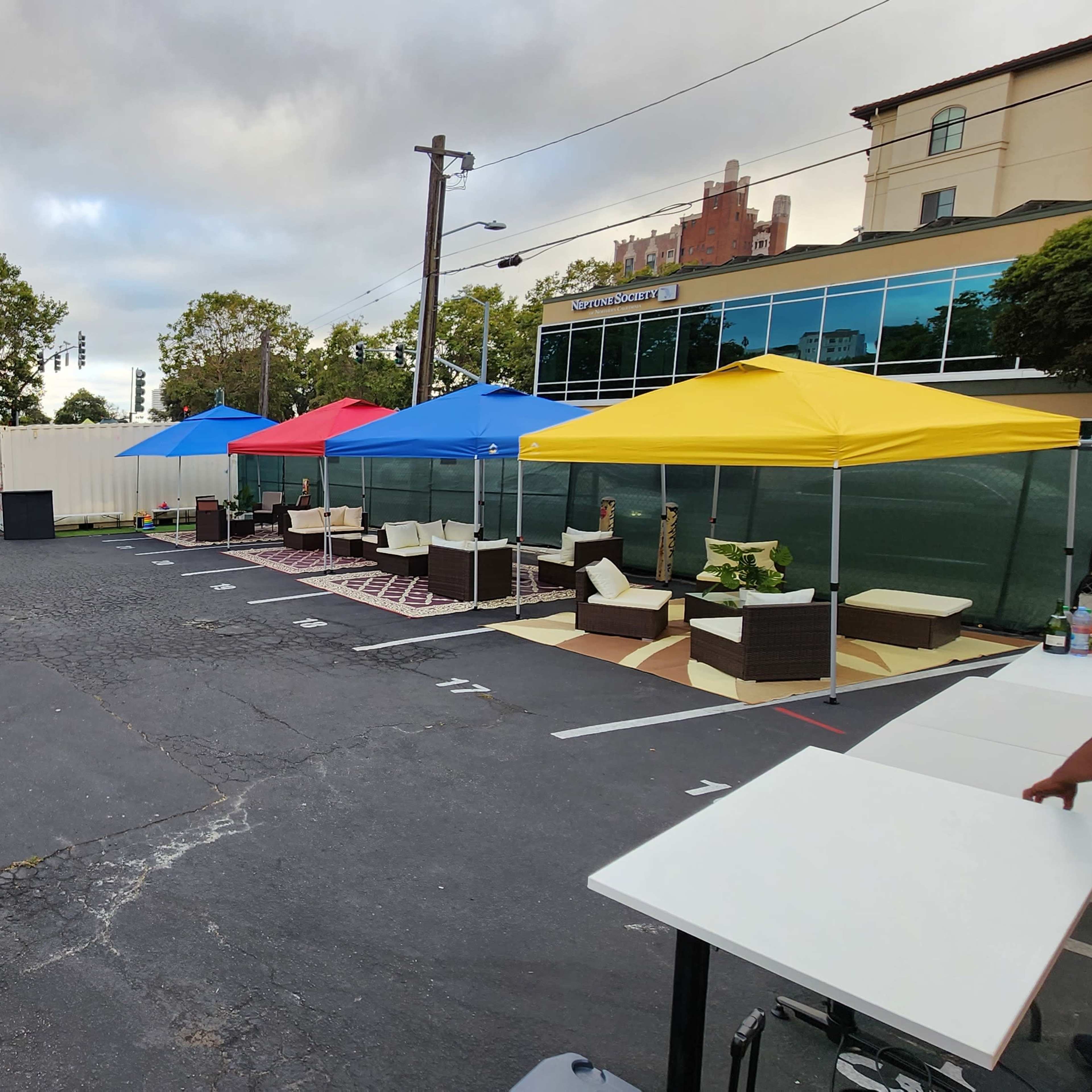 A parking area set up with various colorful tents, seating arrangements, and a backdrop of trees and a building.