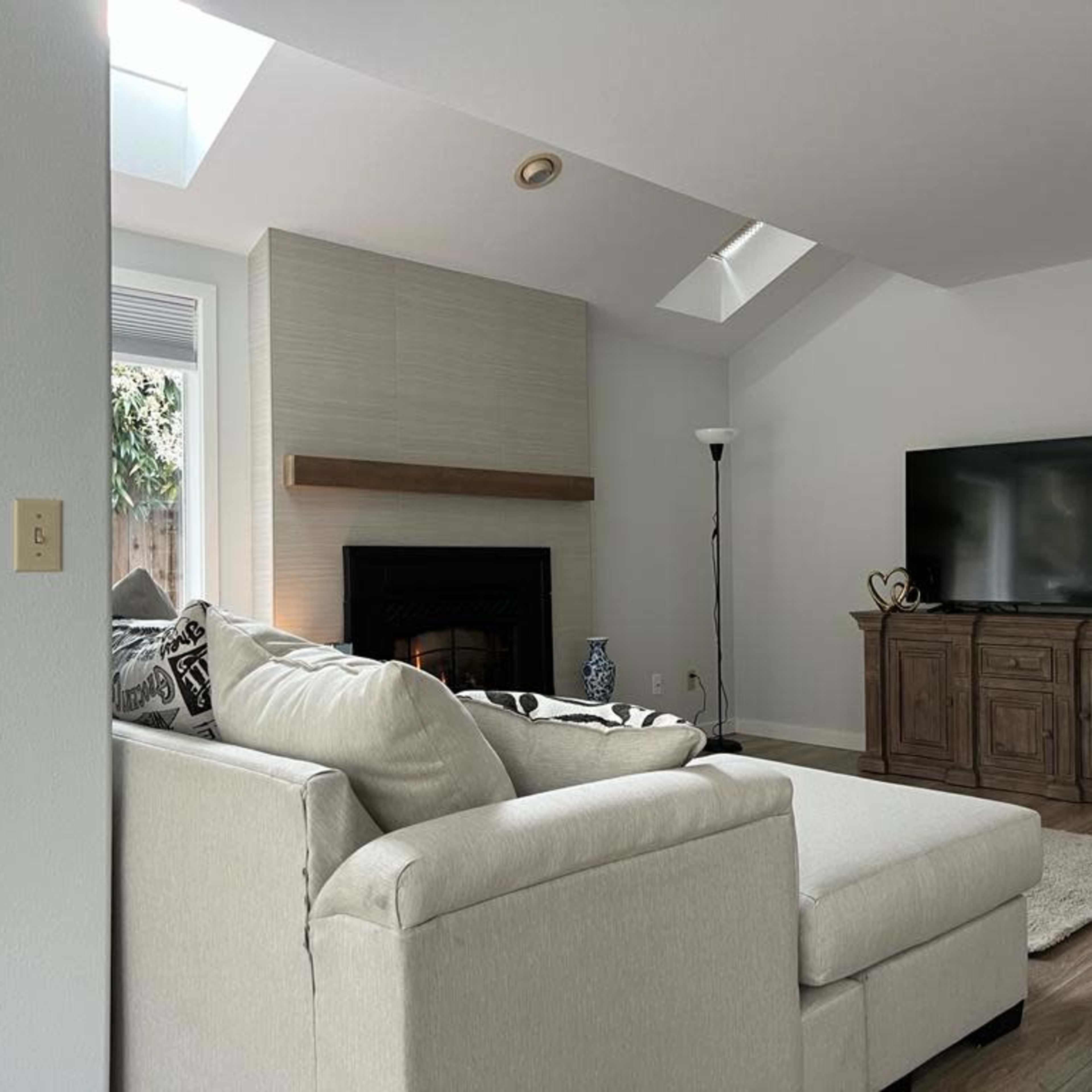 A modern living room with a sectional sofa, a wall-mounted television, and a fireplace underneath a slanted ceiling with skylights.