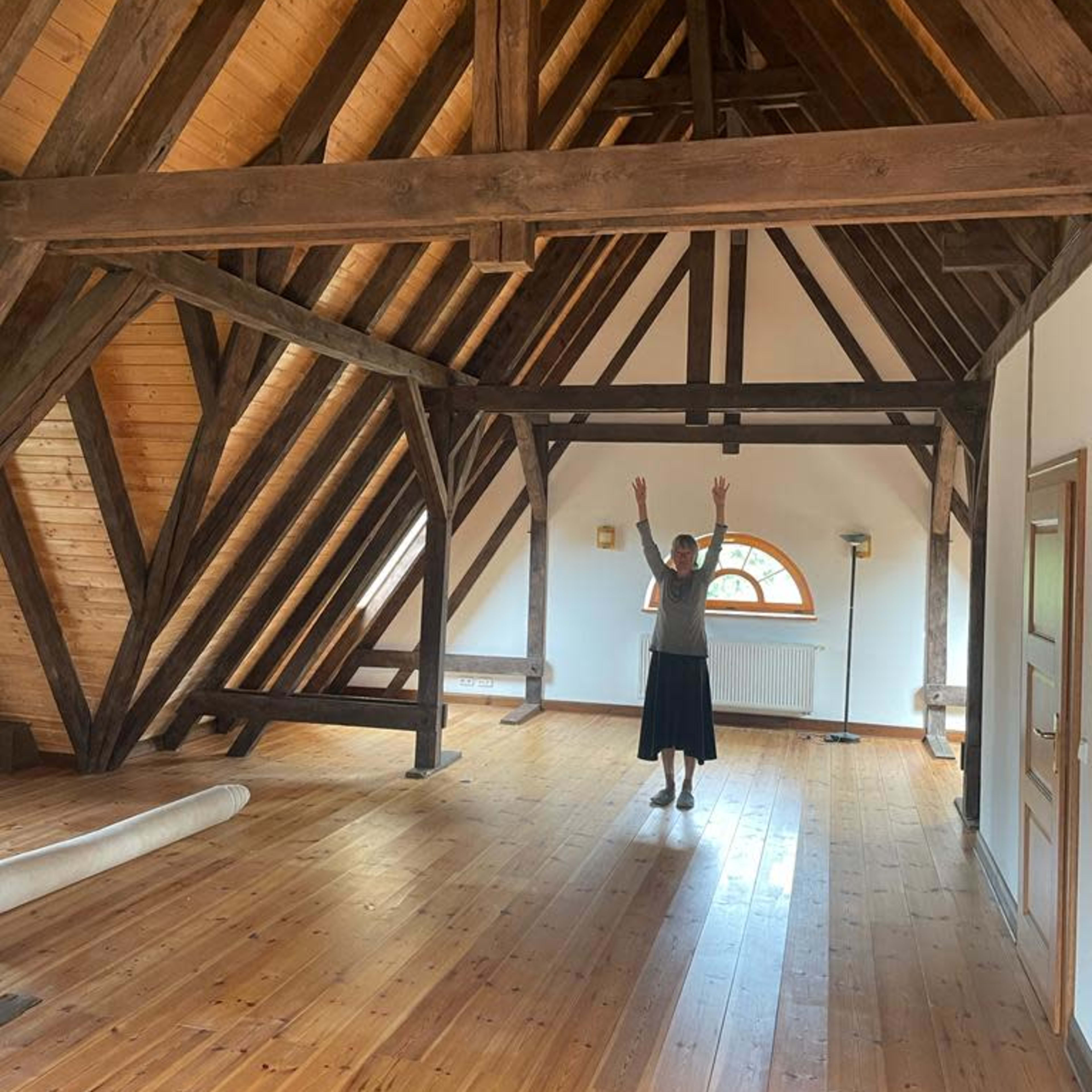 A person stands with arms raised in the center of a spacious room with wooden beams and a hardwood floor.