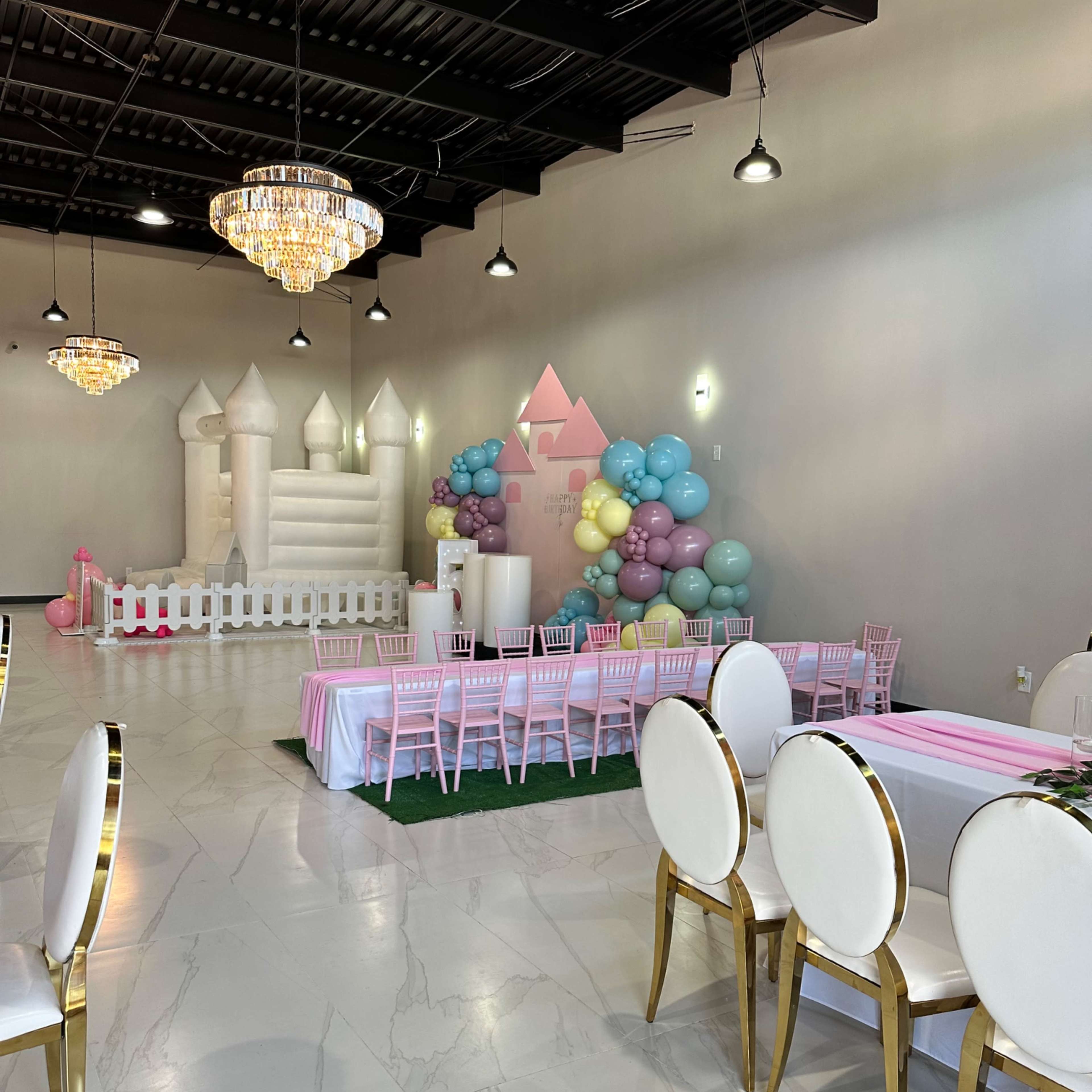An indoor event space decorated with a balloon arch, a castle-shaped play structure, and tables set with pink and white tablecloths.