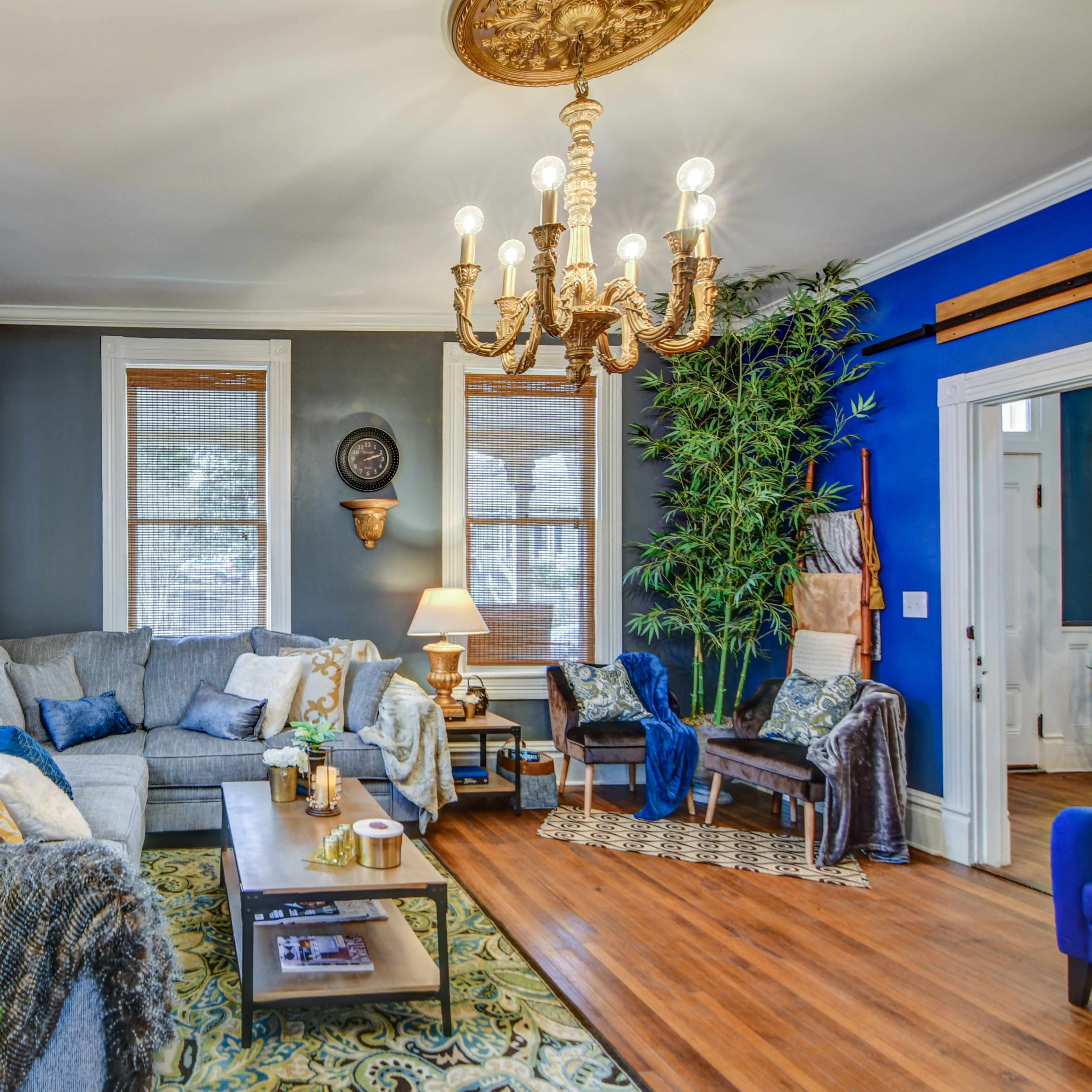 A spacious living room features a large gray sofa, wooden coffee table, and a chandelier, with blue and neutral walls and natural light from the windows.