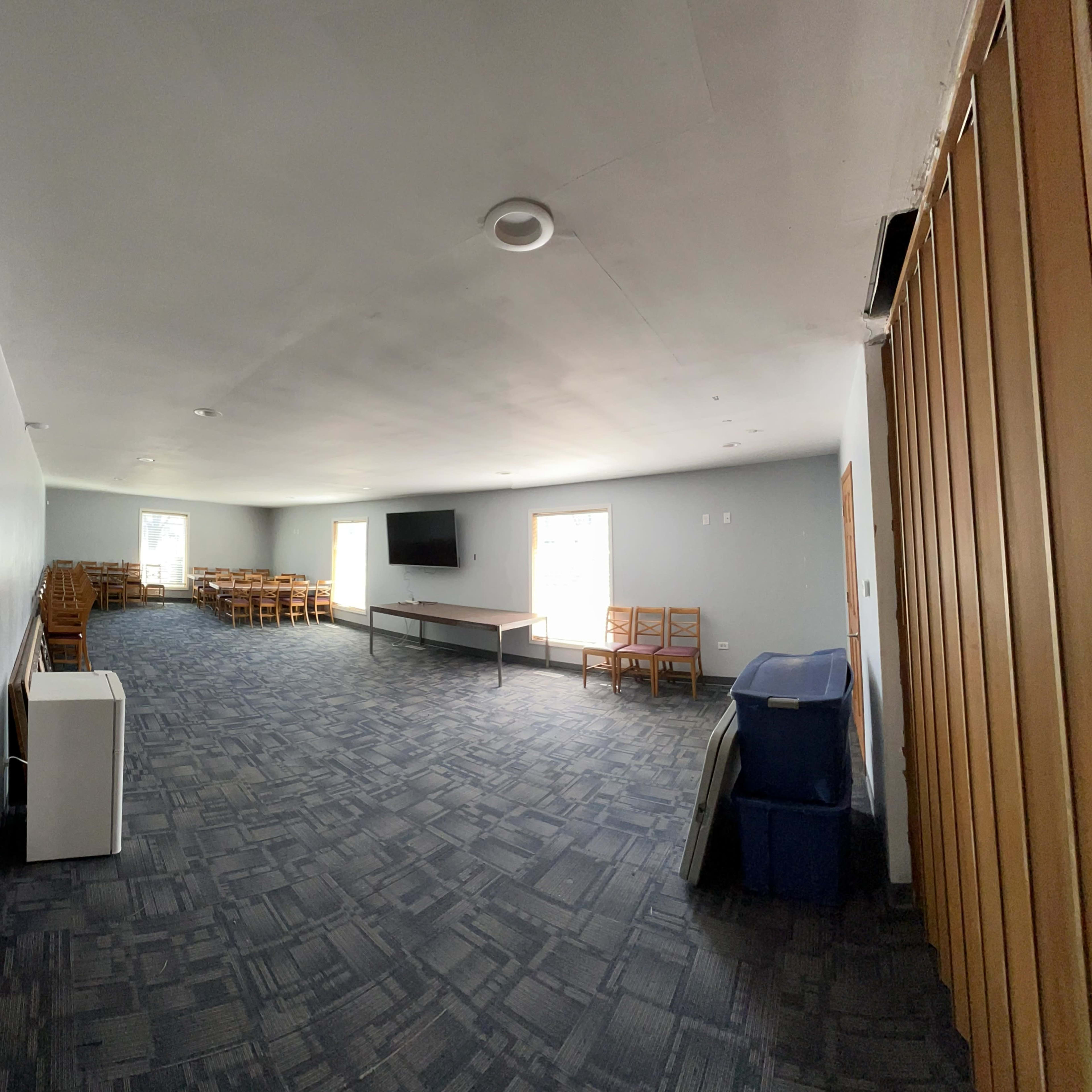 A spacious, empty room with a television mounted on the wall, tables and chairs arranged in the background, and a blue trash bin in the corner.