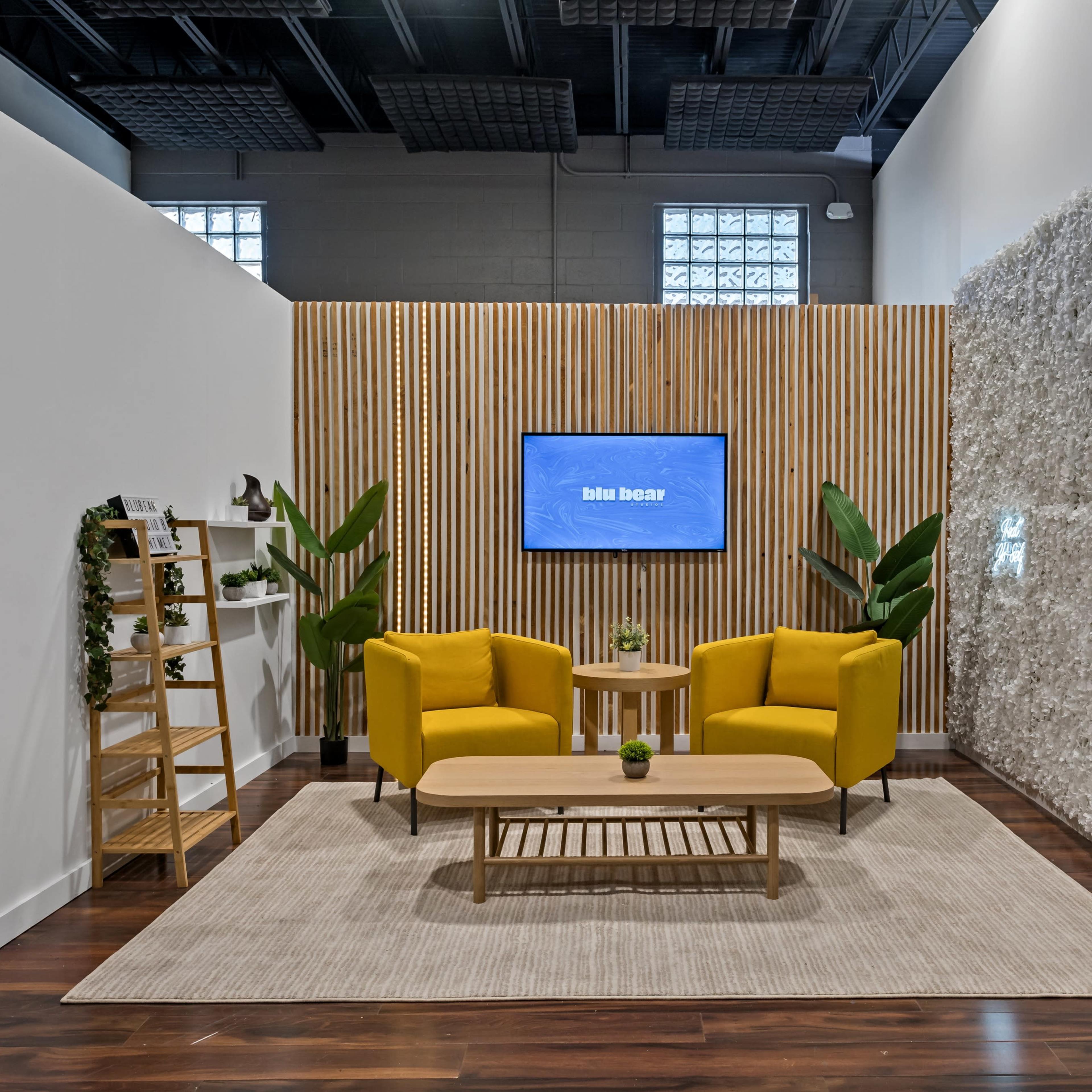 A modern seating area features two yellow chairs and a wooden table on a rug, flanked by a decorative wall and indoor plants, with a television mounted on a wooden backdrop.