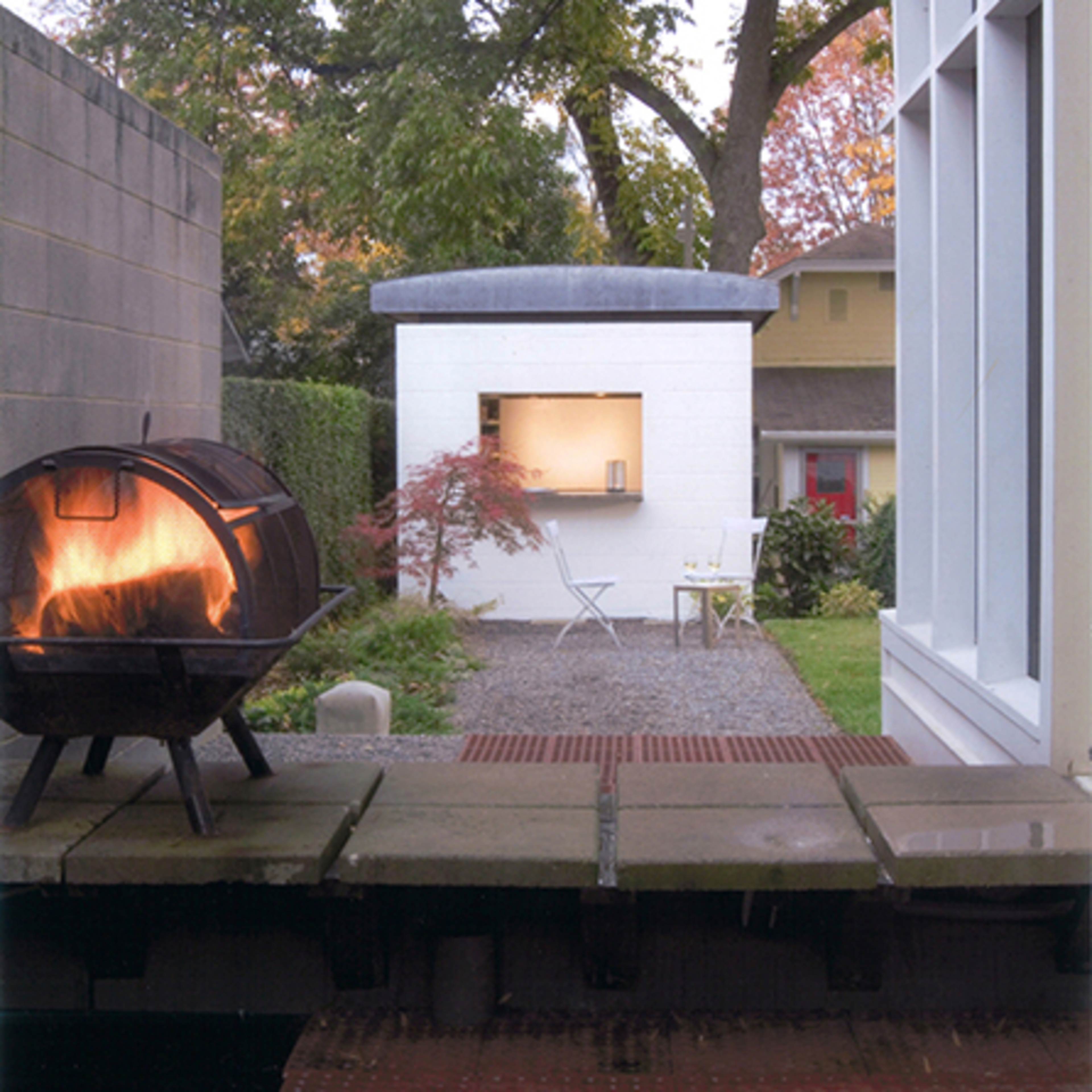 A modern, white shed stands in a backyard next to a stone walkway, with a barrel-shaped fire pit in the foreground and green trees in the background.