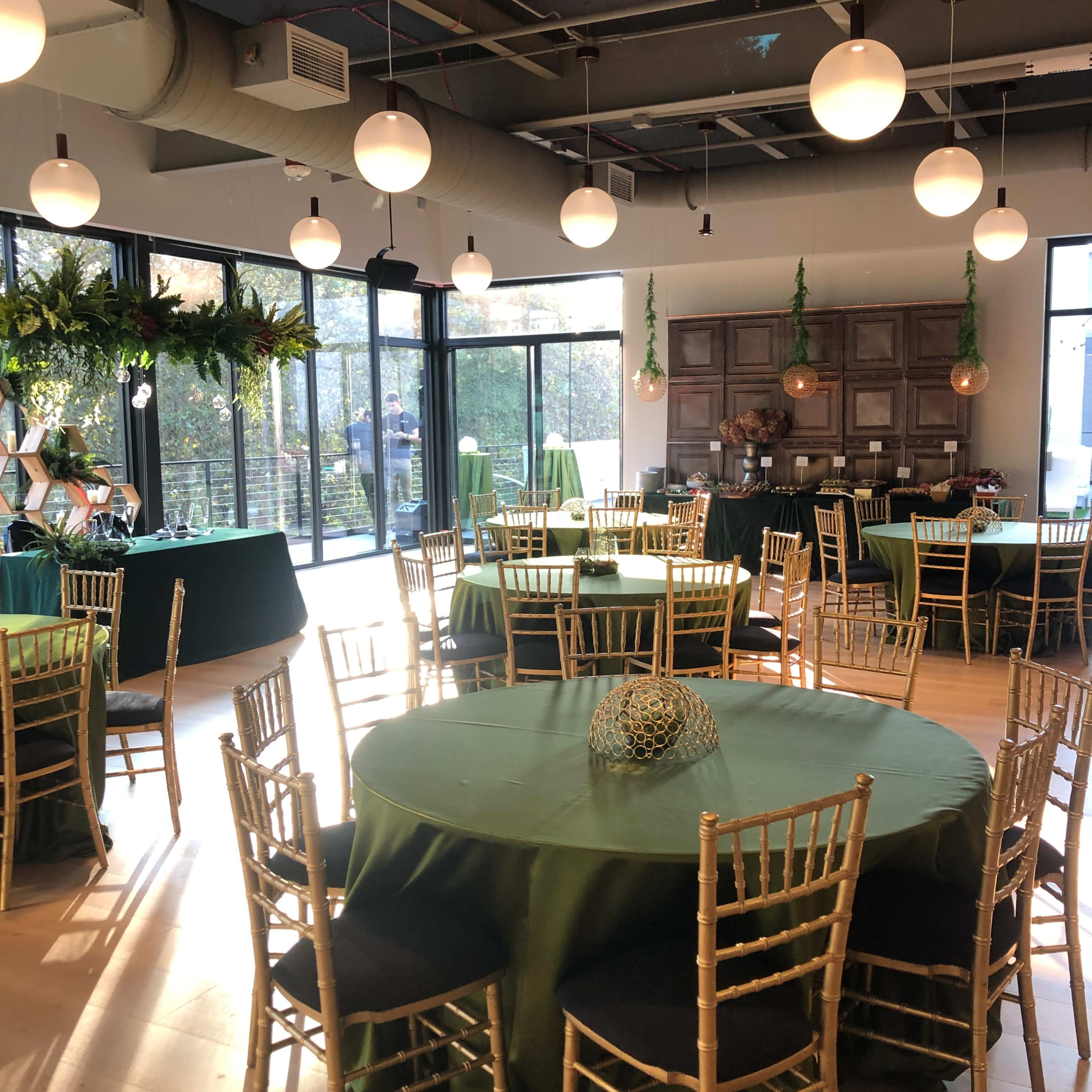 A well-lit event space with several round tables covered in green tablecloths, gold chairs, and decorative elements, along with large windows that provide an outdoor view.