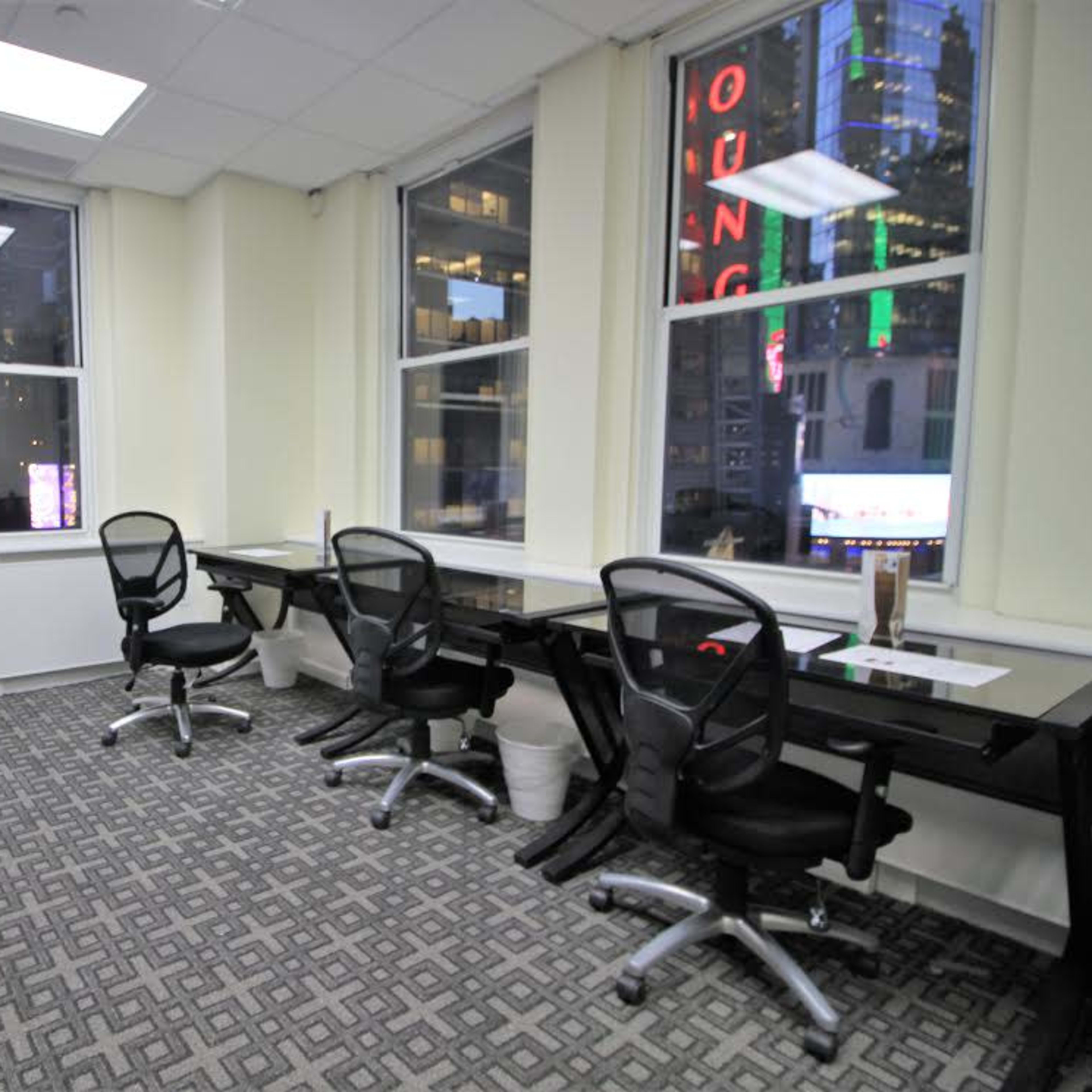 An office space with several empty desks and chairs, illuminated by natural light from large windows.
