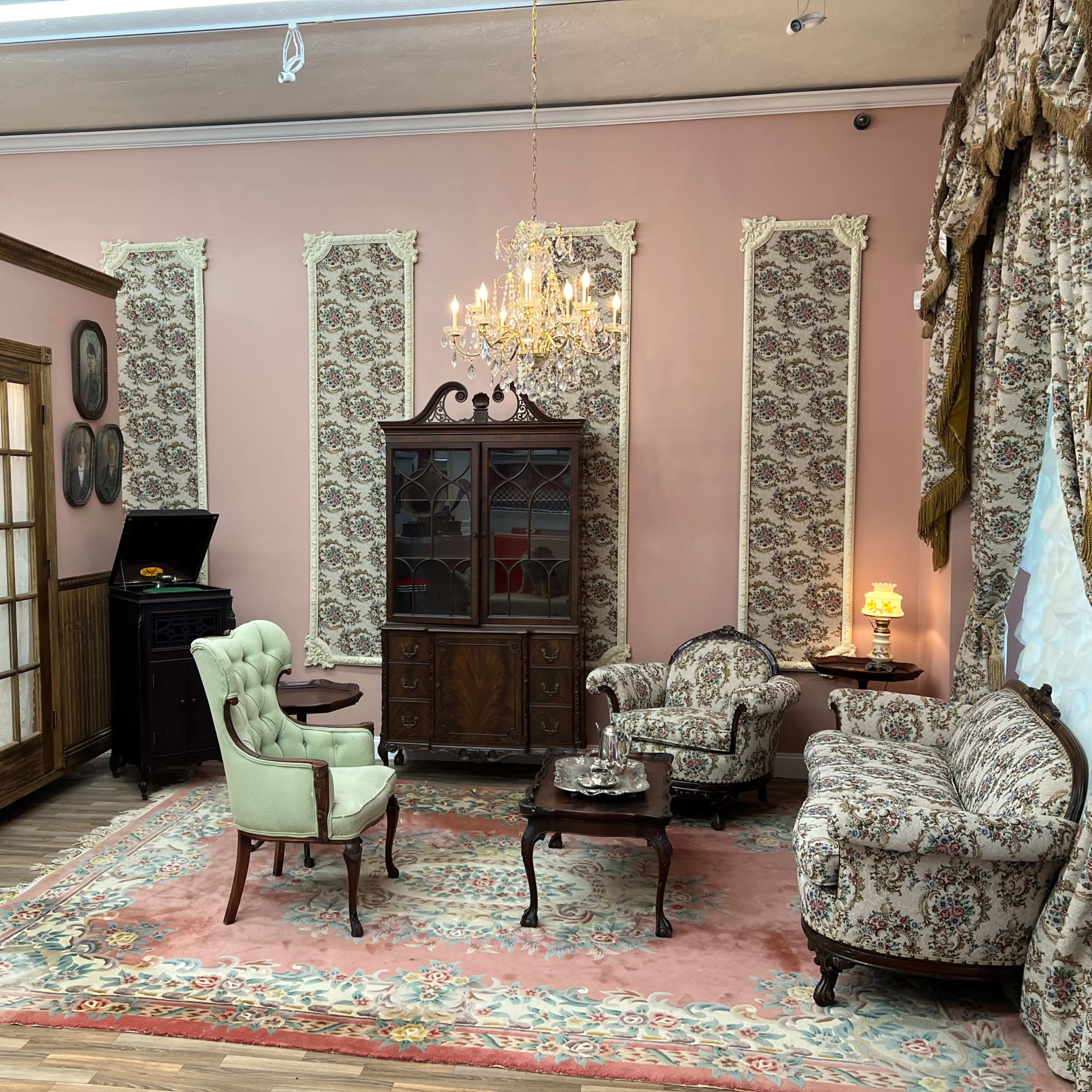 The room features vintage furniture including a chandelier, a patterned area rug, and floral wallpaper.
