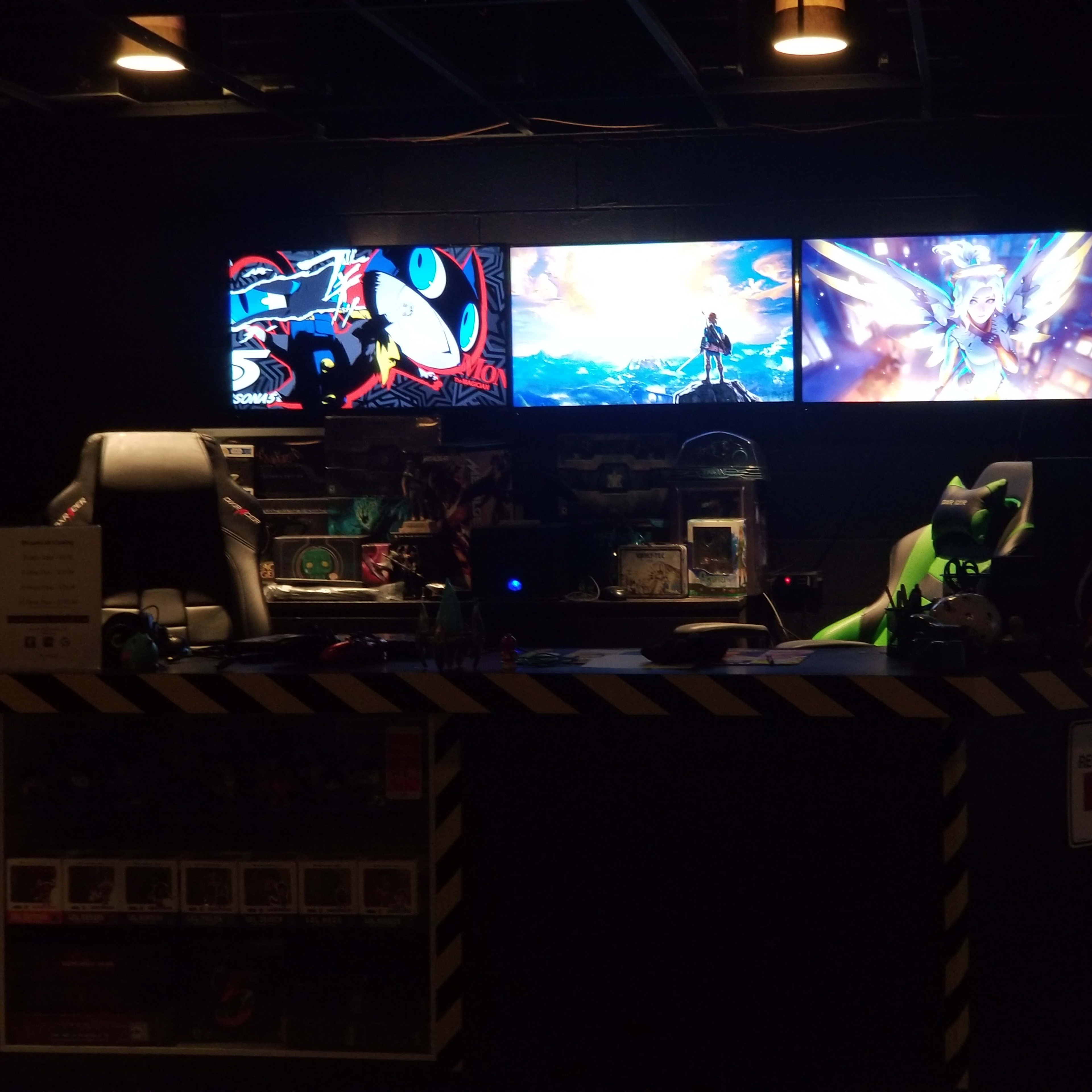 The scene shows a gaming setup with three monitor displays featuring colorful artwork, positioned behind a dark counter with gaming accessories and two gaming chairs.