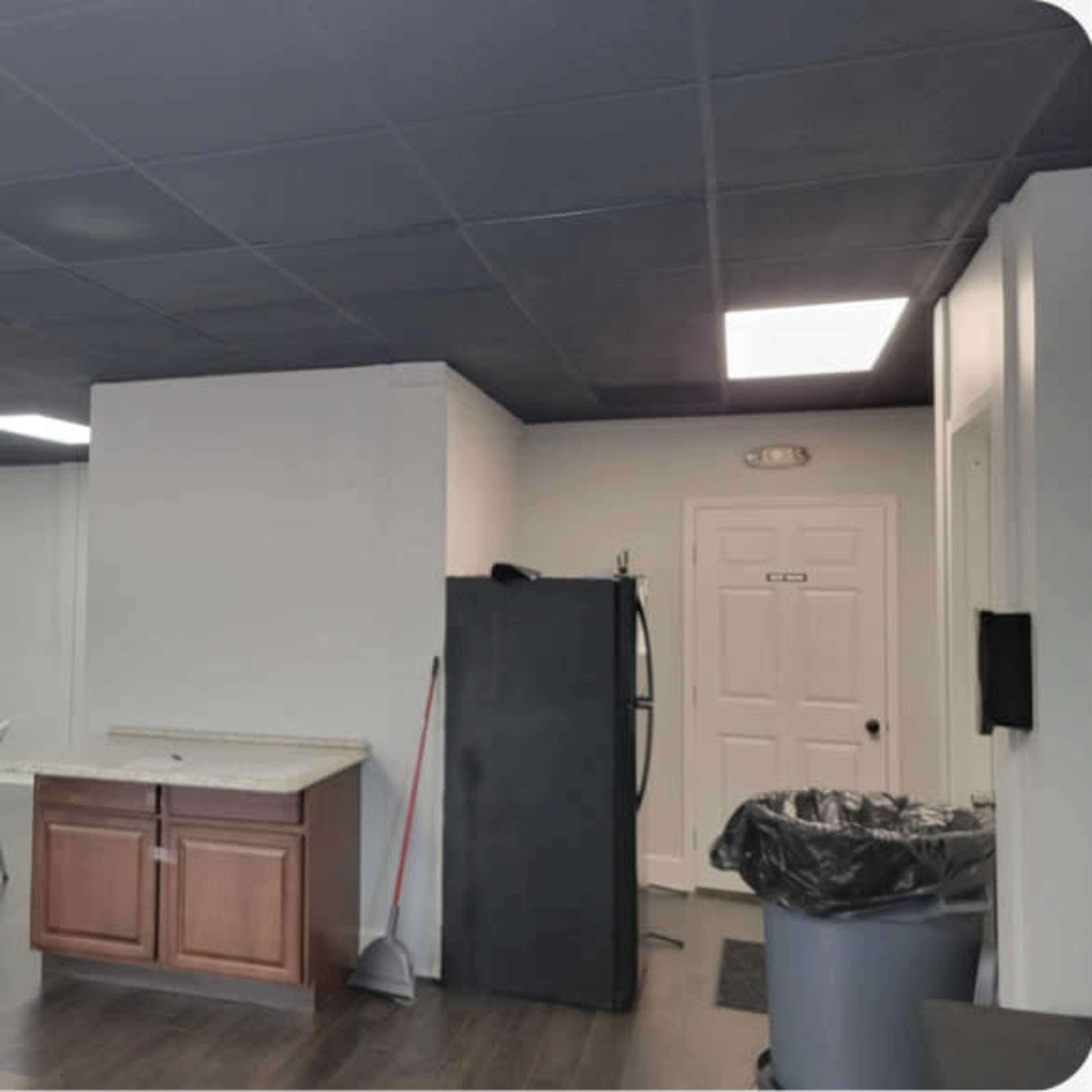 An interior room with a black ceiling, a white wall with a door, a kitchenette area featuring a refrigerator and a sink, and several white folding chairs arranged at a long table.