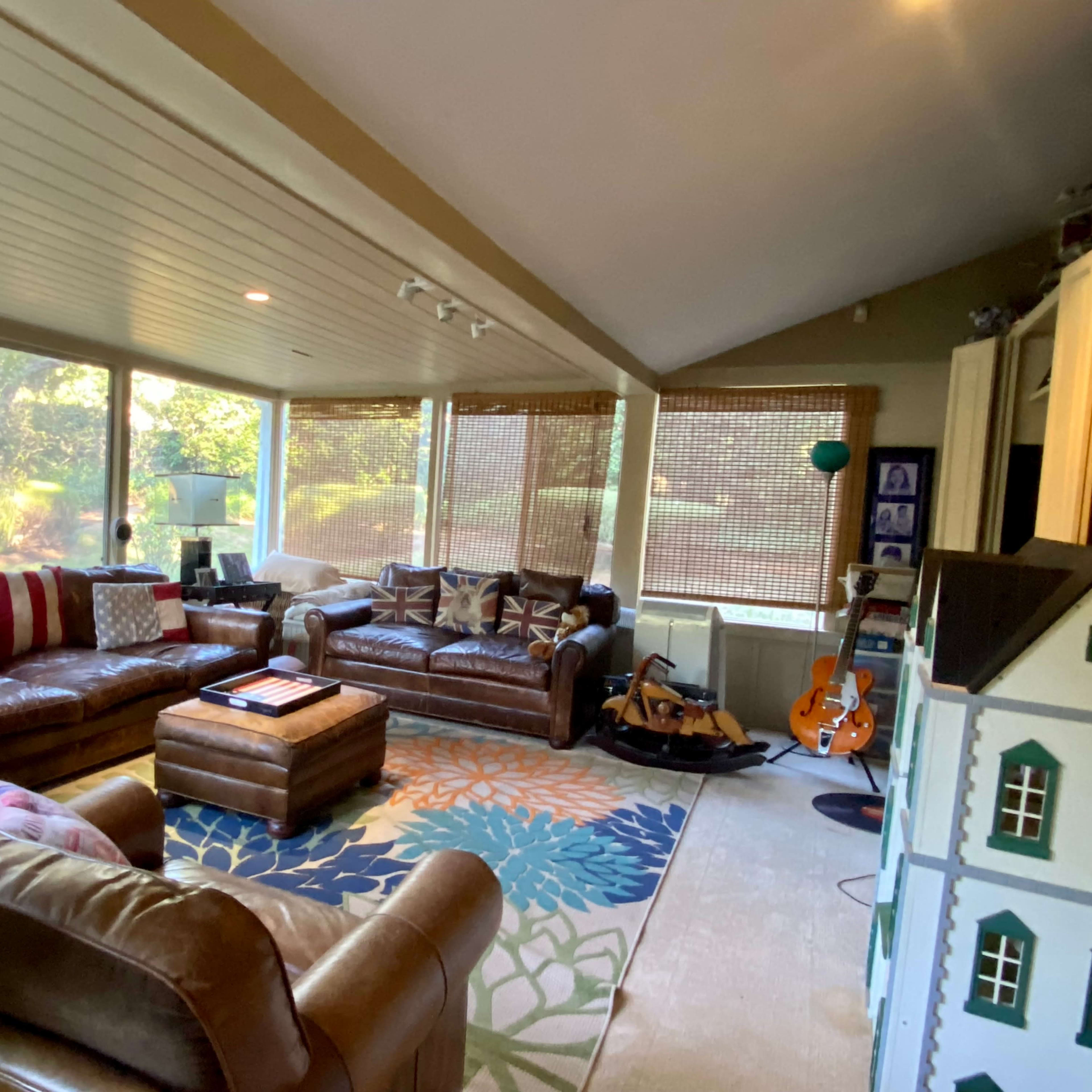 A spacious living room with large windows, a variety of seating options including brown leather sofas, and a colorful area rug.